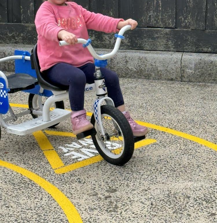 Child riding a blue and white adaptive tricycle on a concrete pavement near a wooden fence, wearing pink shoes, navy leggings, and a pink sweatshirt.
