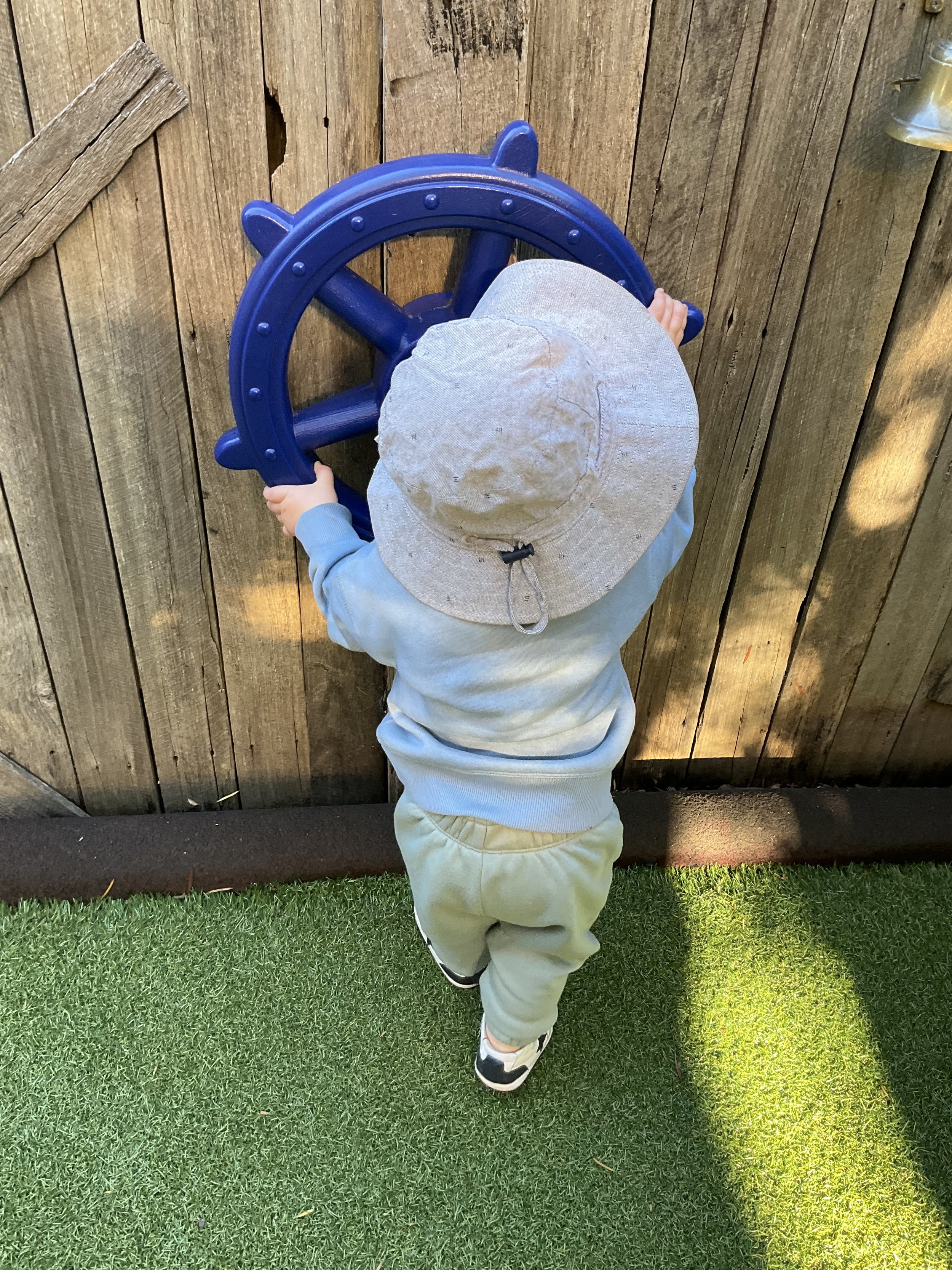 A young child wearing a large sun hat, a light blue long sleeve shirt, beige pants, and white sneakers with black accents, is standing outdoors against a wooden fence and green grass, holding onto a blue ship wheel.