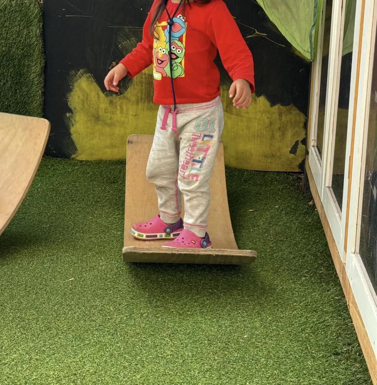 Child wearing a red 'Sesame Street' sweatshirt, gray sweatpants with colorful lettering, and pink Crocs standing on a skateboard ramp indoors with artificial green grass flooring and a painted backdrop.
