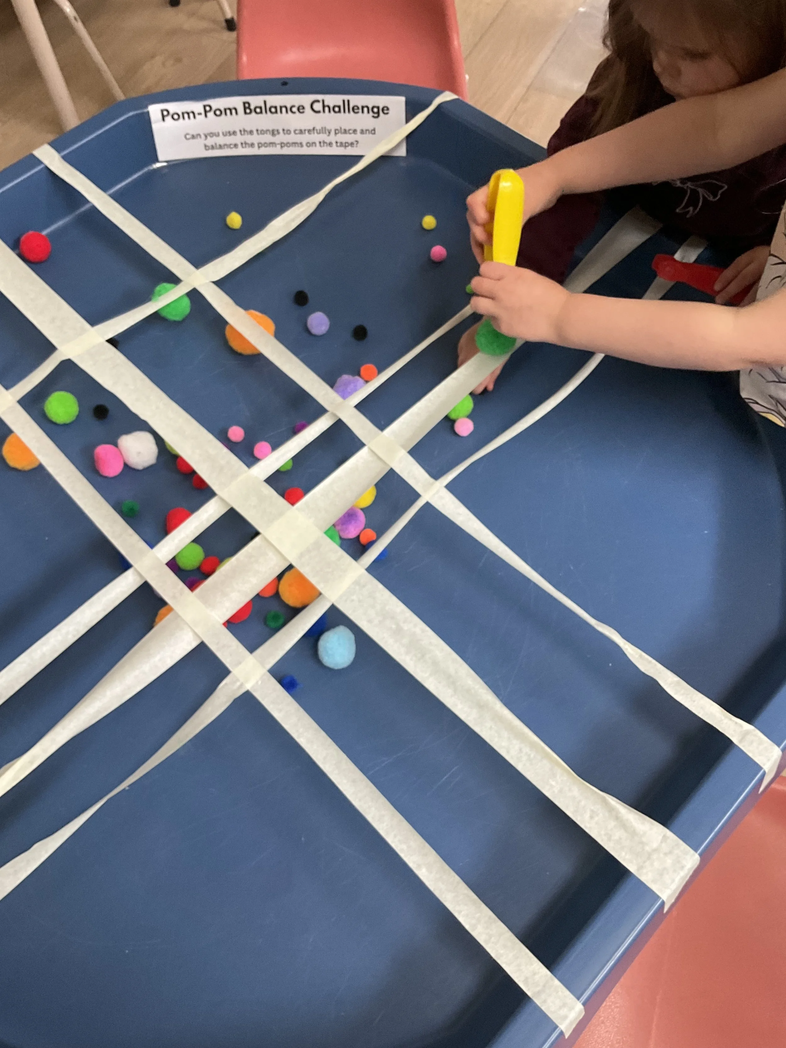 Kids participating in a balance challenge by using tongs to carefully place and balance the pom-poms on the tape. 