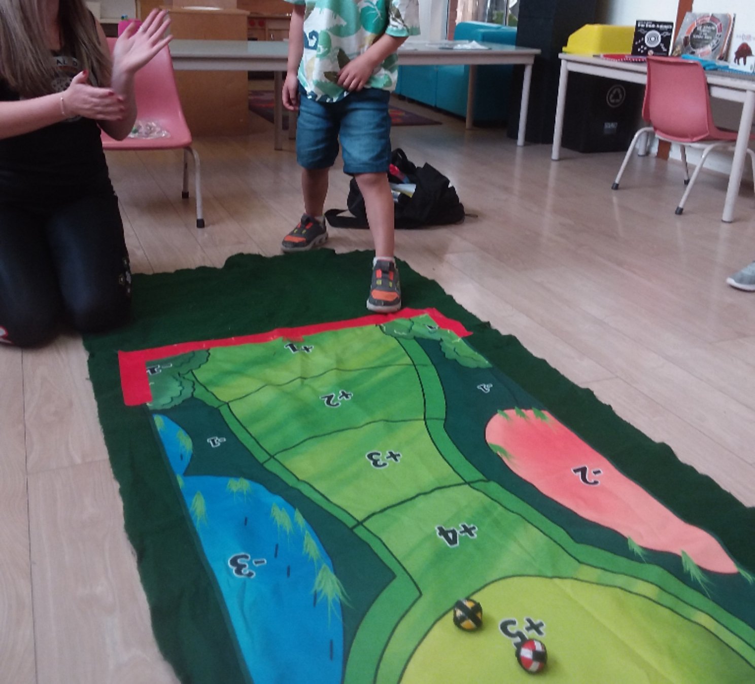 Children playing a board game on the floor featuring a nature-themed path with numbered spaces, trees, water, and hills.