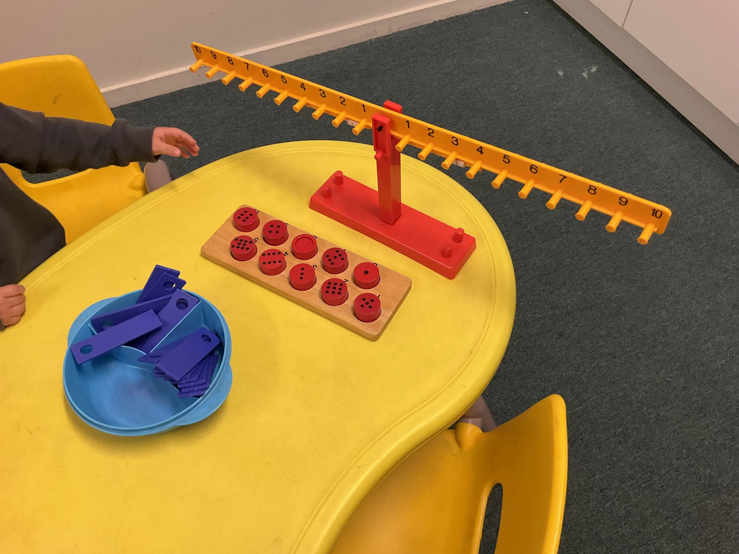 Child reaching for a yellow and red peg game on a yellow table, with blue plastic pieces nearby.