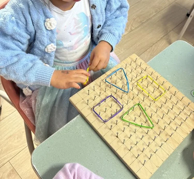 A young girl is working on a shape-matching activity with a wooden pegboard and colorful pipe cleaners, forming various geometric shapes like a triangle, square, and rectangle.