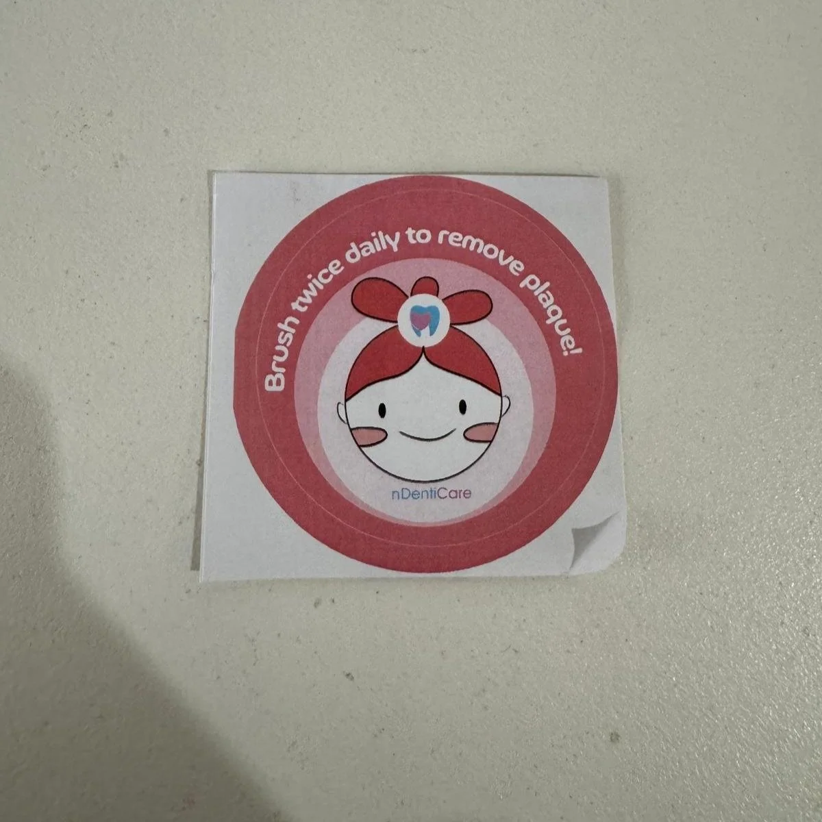 Sticker with a smiling cartoon girl and pink hair tied in a bow, surrounded by a red circle with white text that reads 'Brush twice daily to remove plaque!'