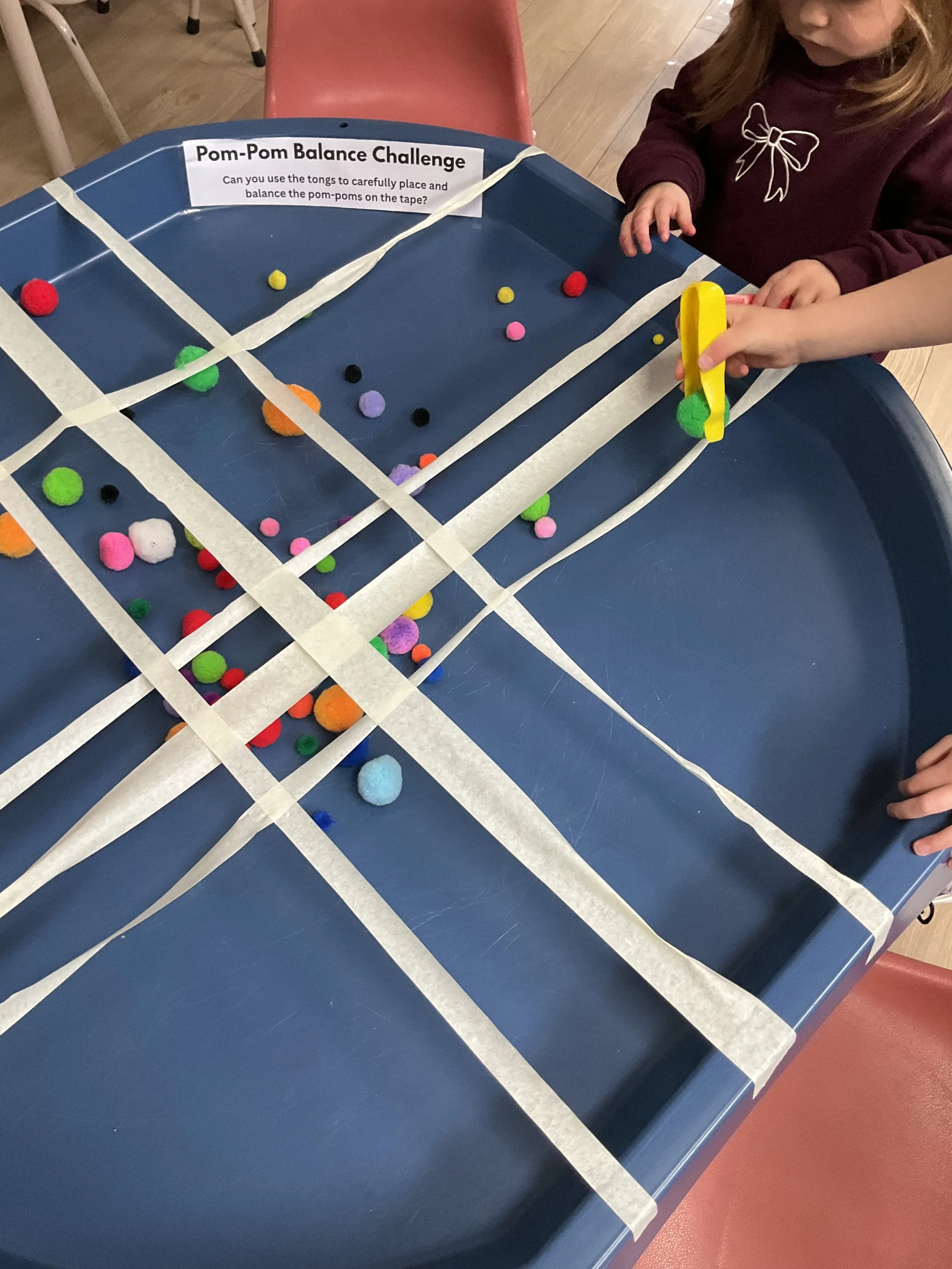 A child participating in a Pom-Pom Balance Challenge, using tongs to carefully place pom-poms on tape on a blue tray, with a sign displaying instructions.