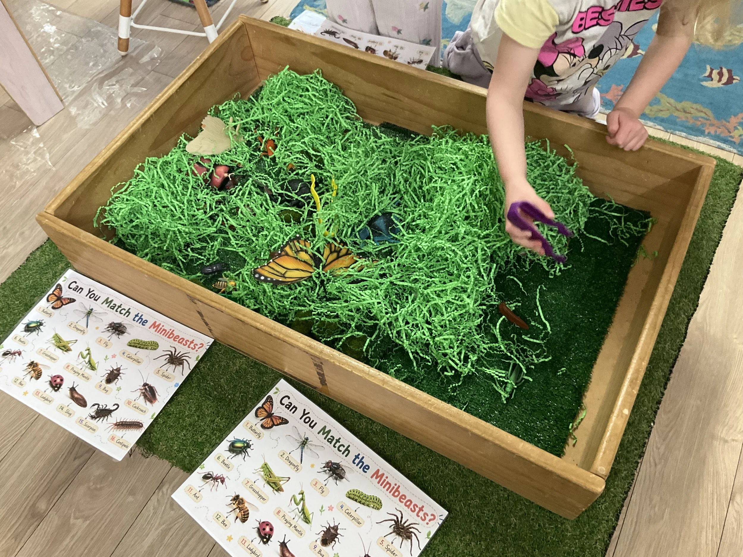 A display table with green shredded paper grass filled with toy bugs, insects, and spiders. Two children are interacting with the display, one reaching in with purple tongs.