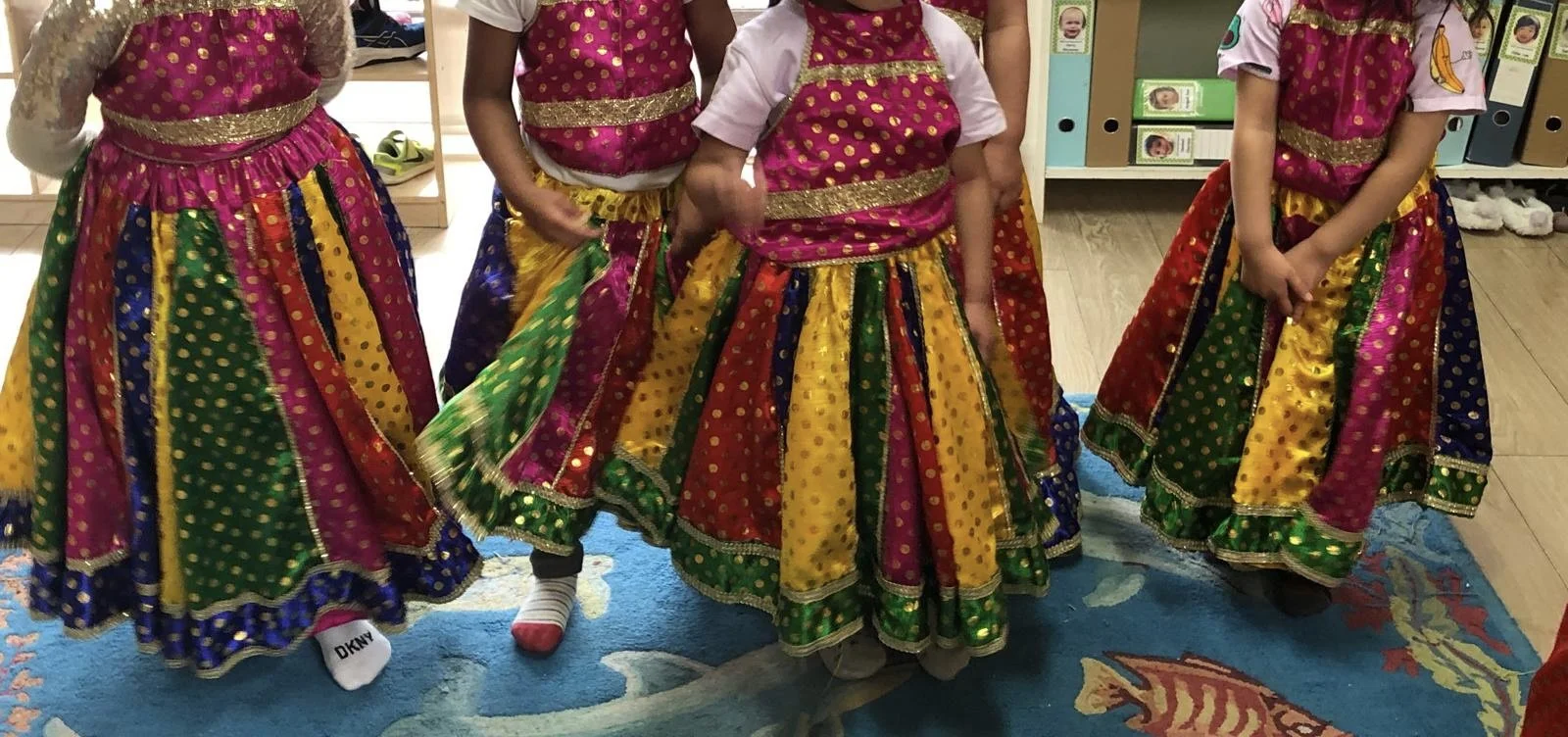 Children wearing colorful traditional Indian attire, including skirts with bright colors and gold embellishments, standing together indoors on a rug with a fish design for a Bollywood Dance Incursion.