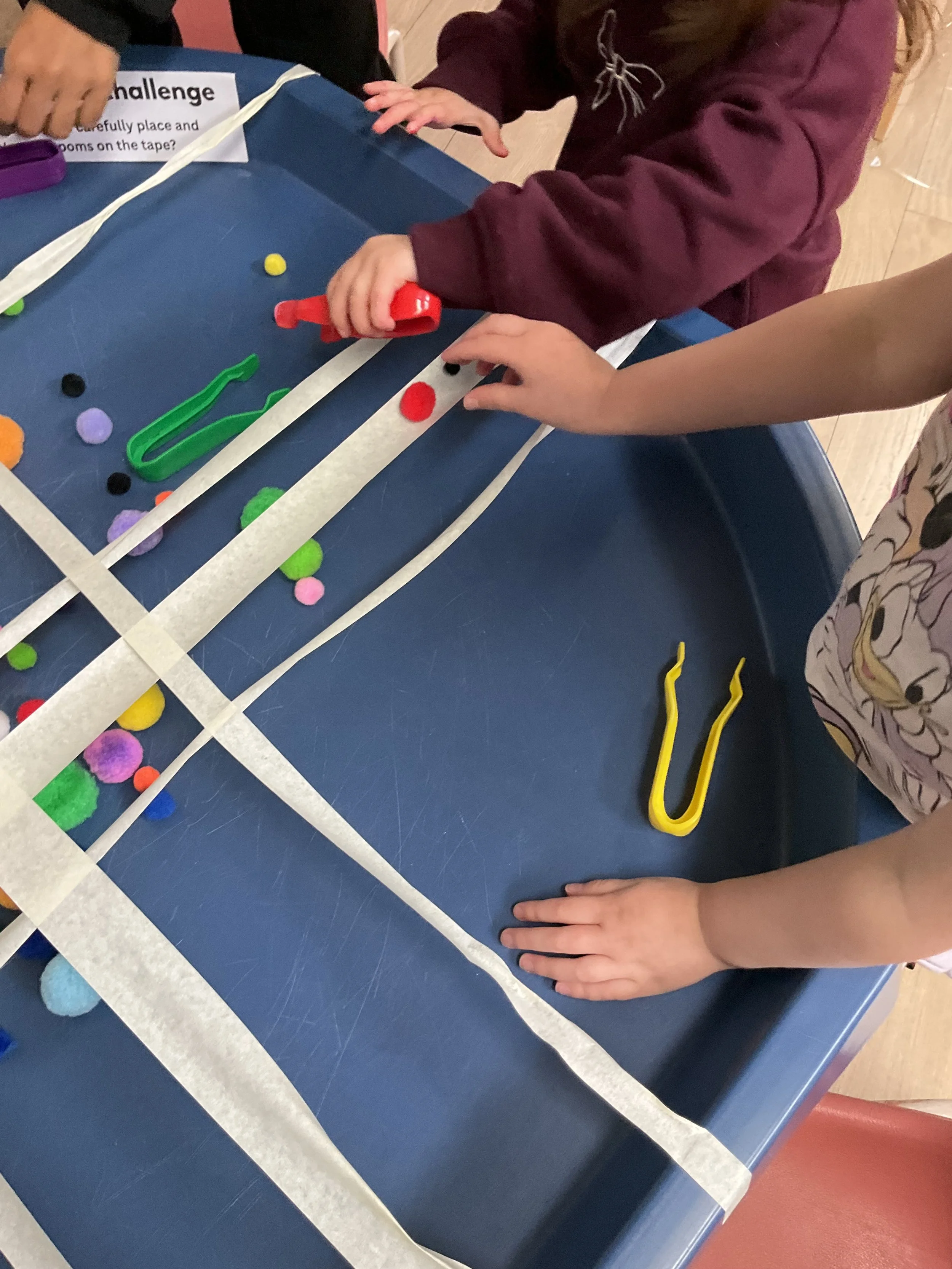 Kids participating in a balance challenge by using tongs to carefully place and balance the pom-poms on the tape. 