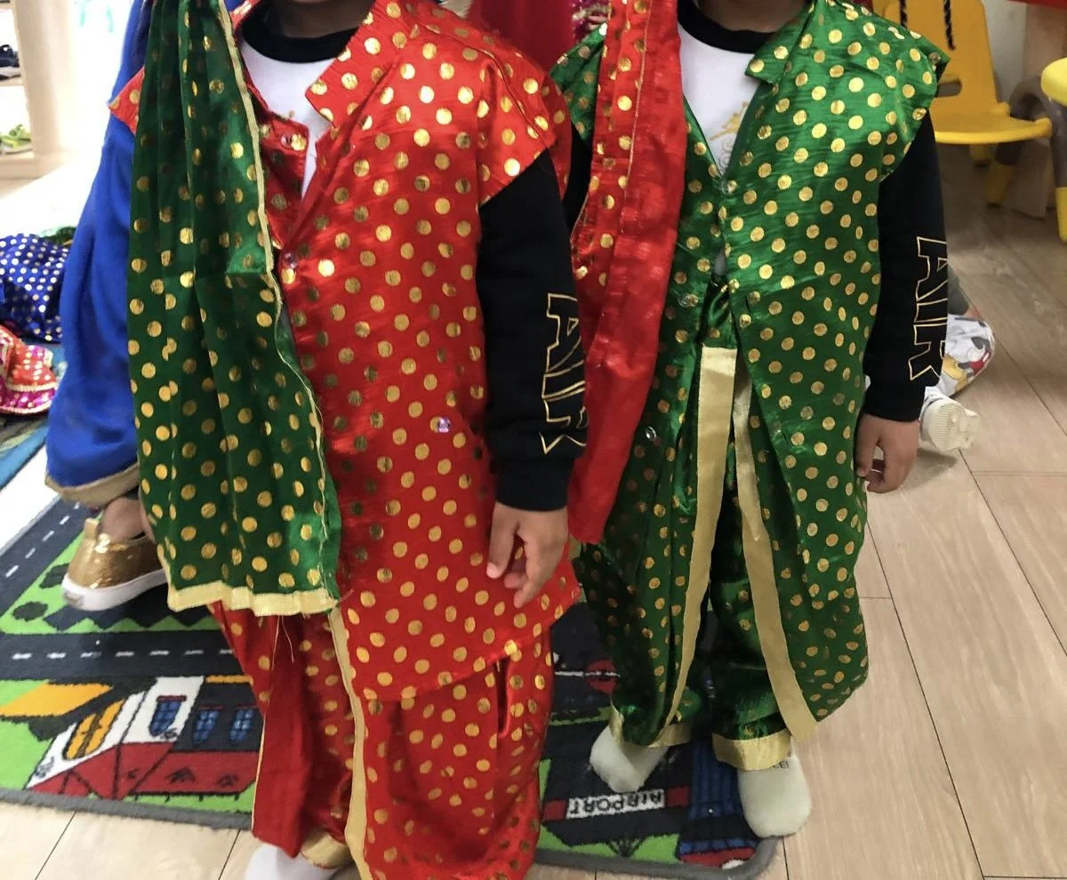 Children dressed in colorful, traditional Asian costumes with polka dot patterns, standing on a play mat with a street and house design.