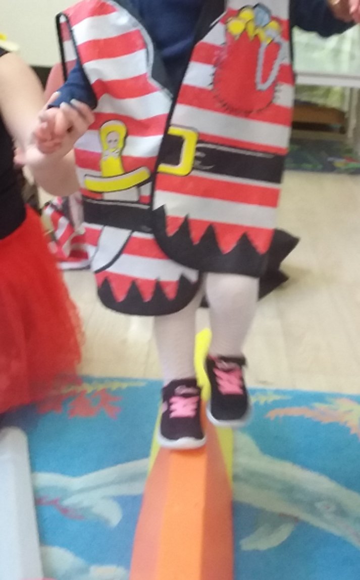 Child balancing on a seesaw in a classroom, dressed in a pirate-themed costume with striped red and white fabric, cartoon graphics, and black shoes with pink accents.