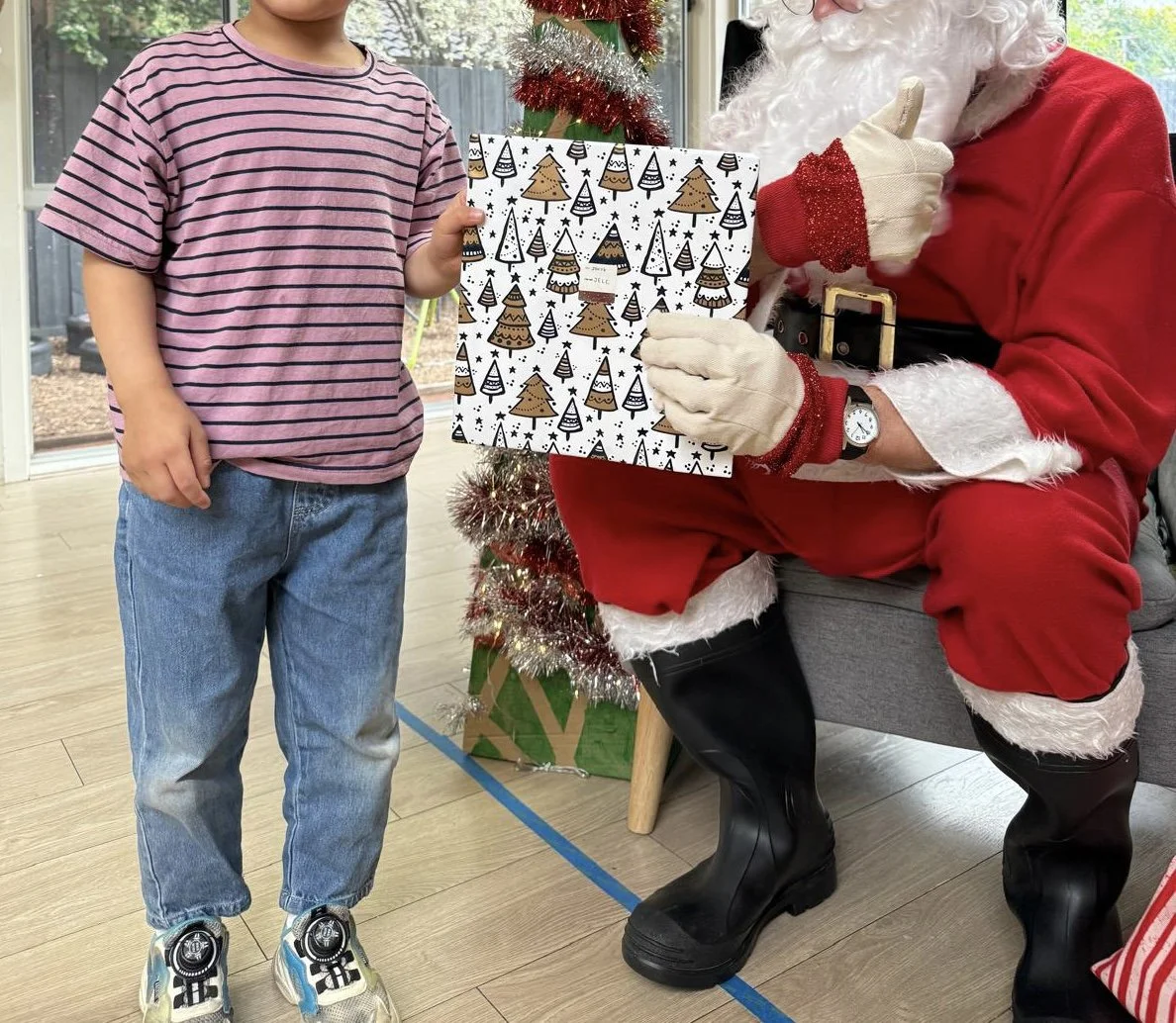 A child in a pink and navy striped shirt and jeans standing next to Santa Claus, who is sitting and holding a Christmas gift with Christmas tree and tinsel decorations in the background.