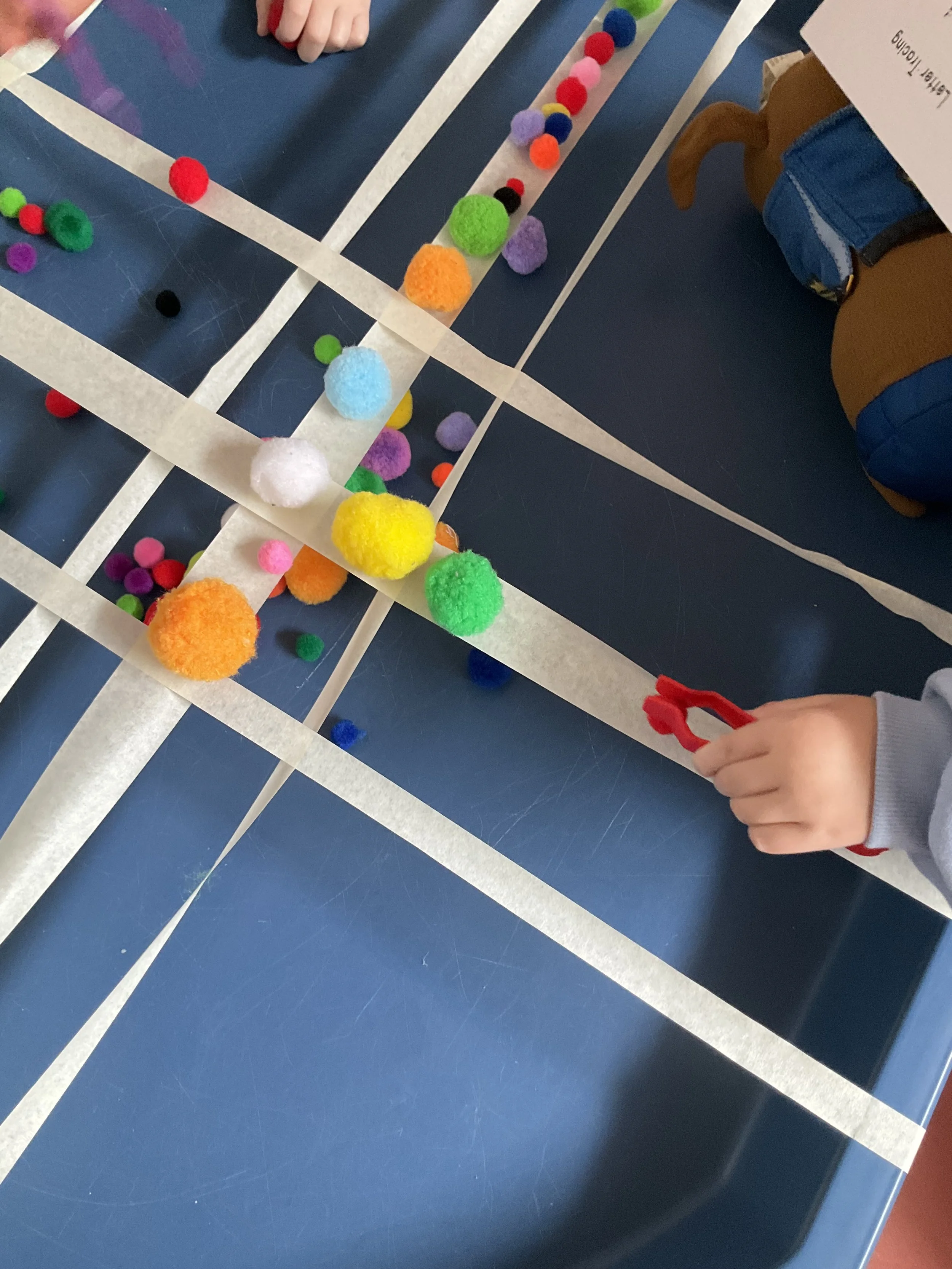 Kids participating in a balance challenge by using tongs to carefully place and balance the pom-poms on the tape.