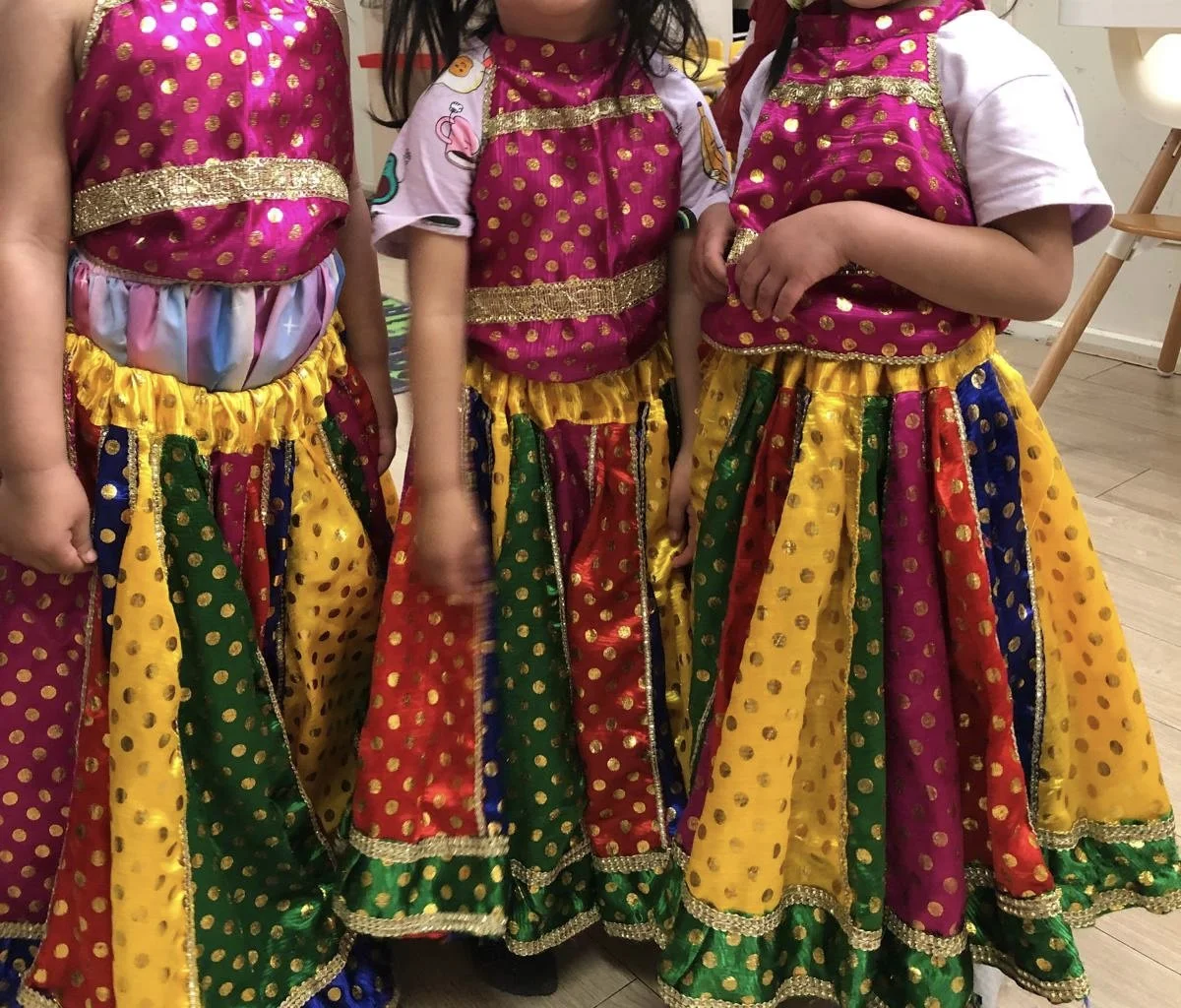 Three children wearing traditional colorful Indian dresses with gold embellishments, featuring pink, yellow, green, and blue fabrics with gold polka dots for a Bollywood Dance Incursion.