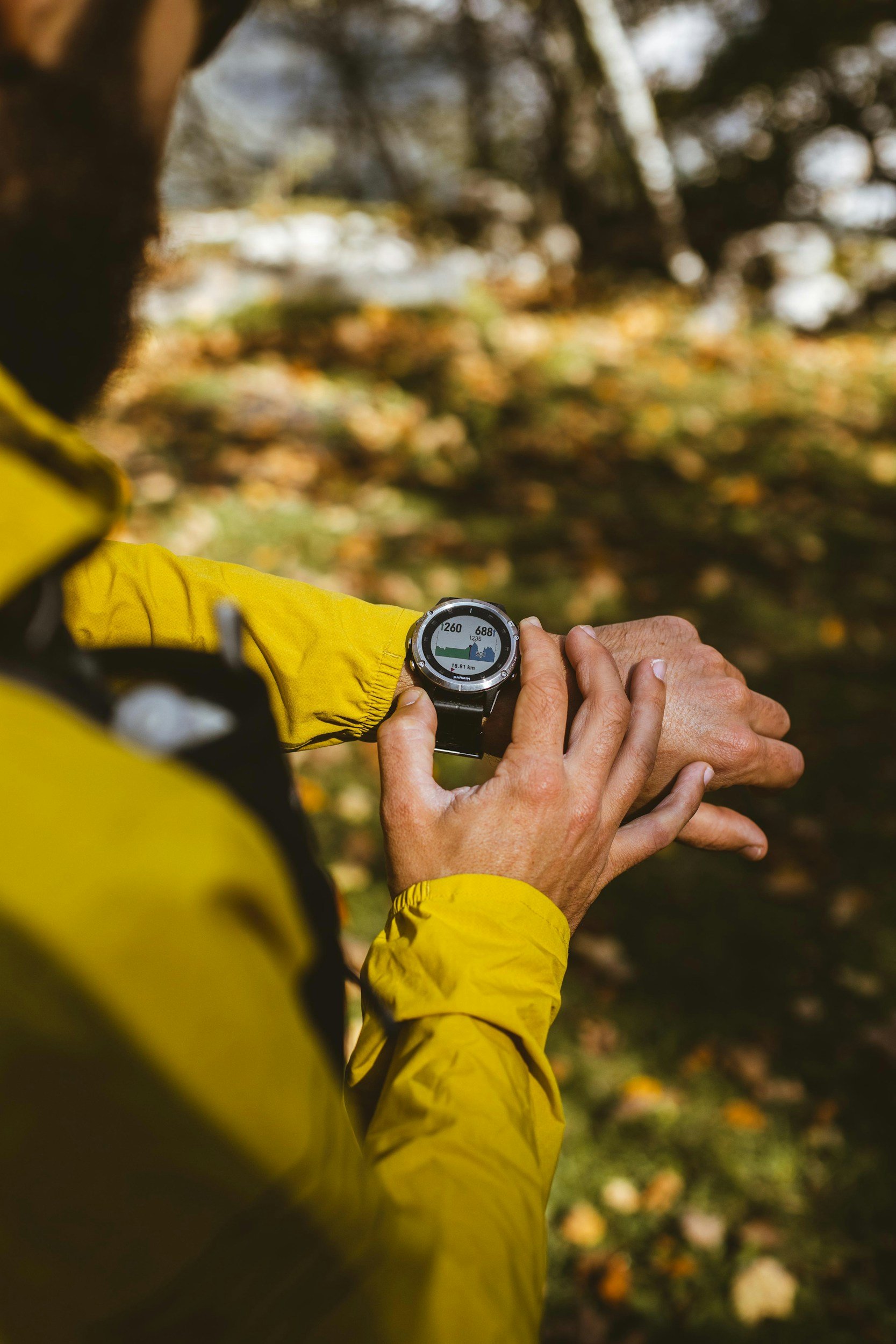 Close-up of a person in a yellow jacket checking a smartwatch outdoors with blurred autumn trees in the background.