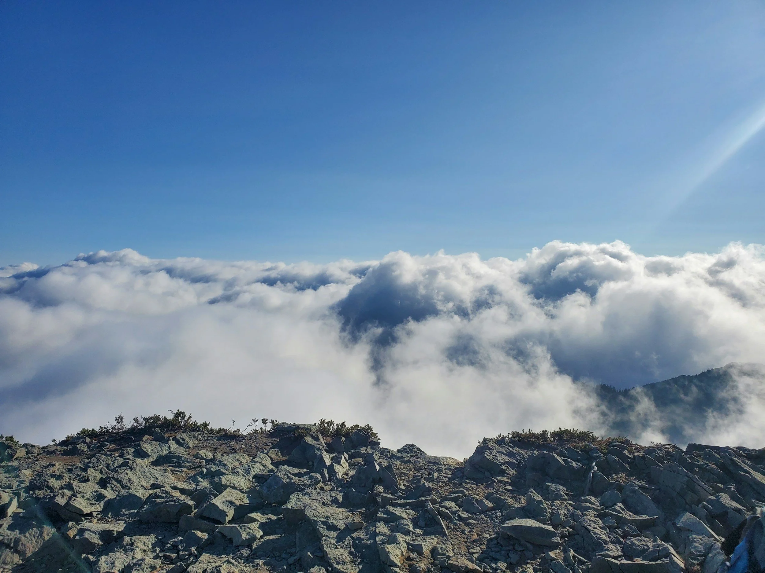 Mountaintop with rocky terrain in the foreground, clouds below, and a clear blue sky above.
