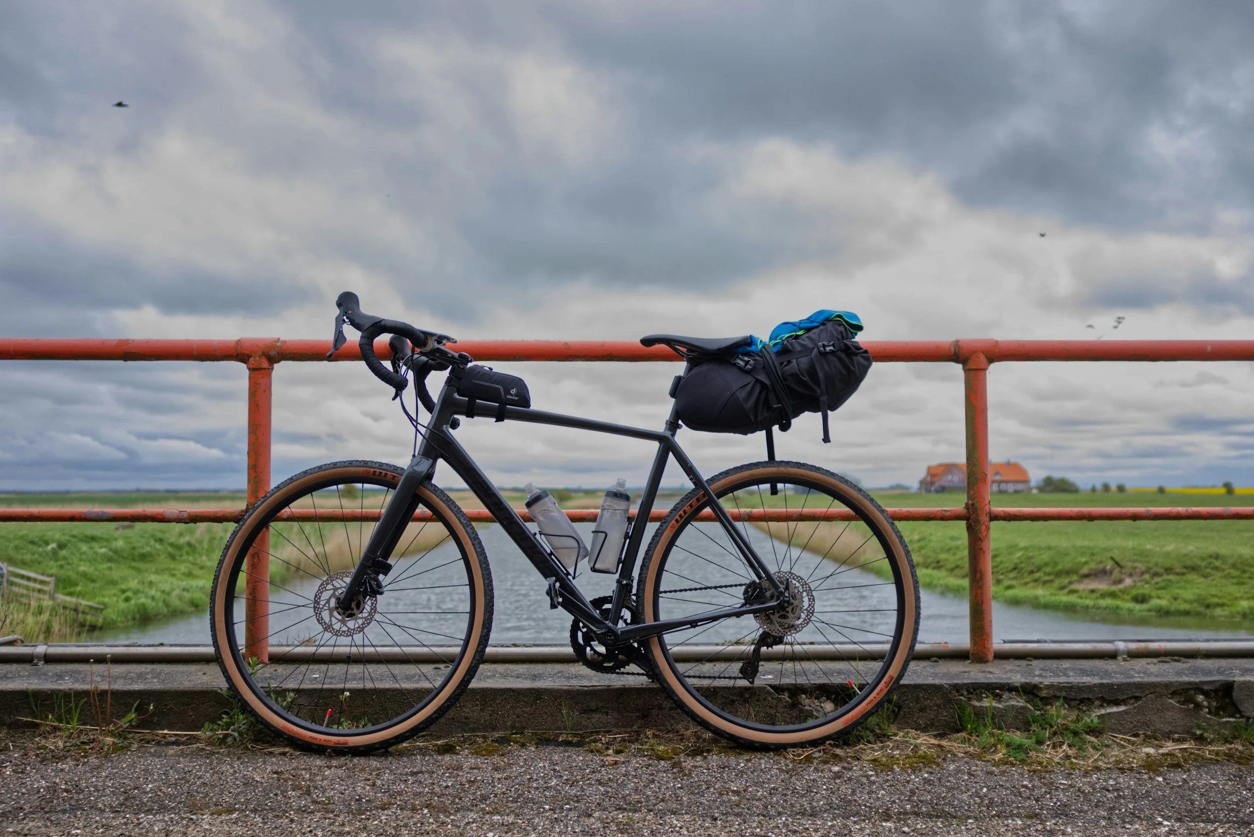 A black bicycle with a rear bag and water bottles mounted on the frame, leaning against a red metal railing, with a cloudy sky, a house, and open fields in the background.