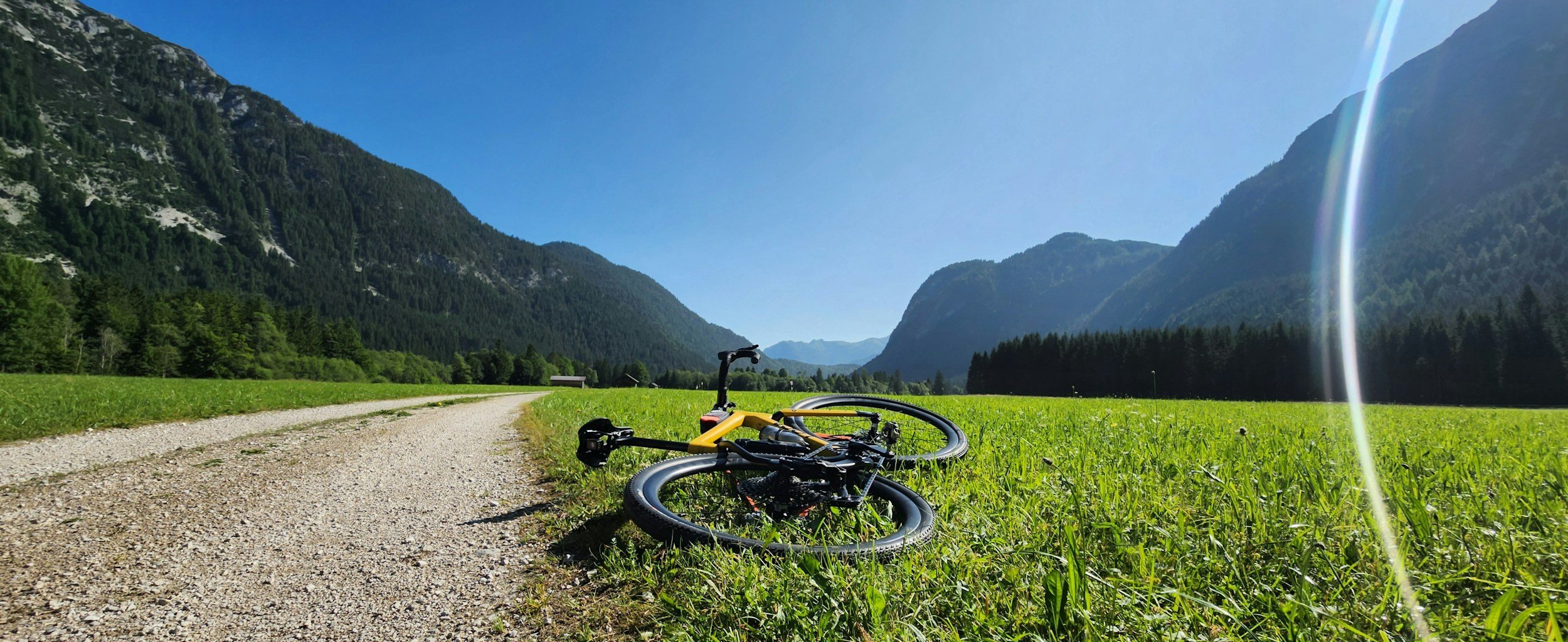 A mountain bike lying on a grassy field beside a gravel path, with tall mountains and a clear blue sky in the background.