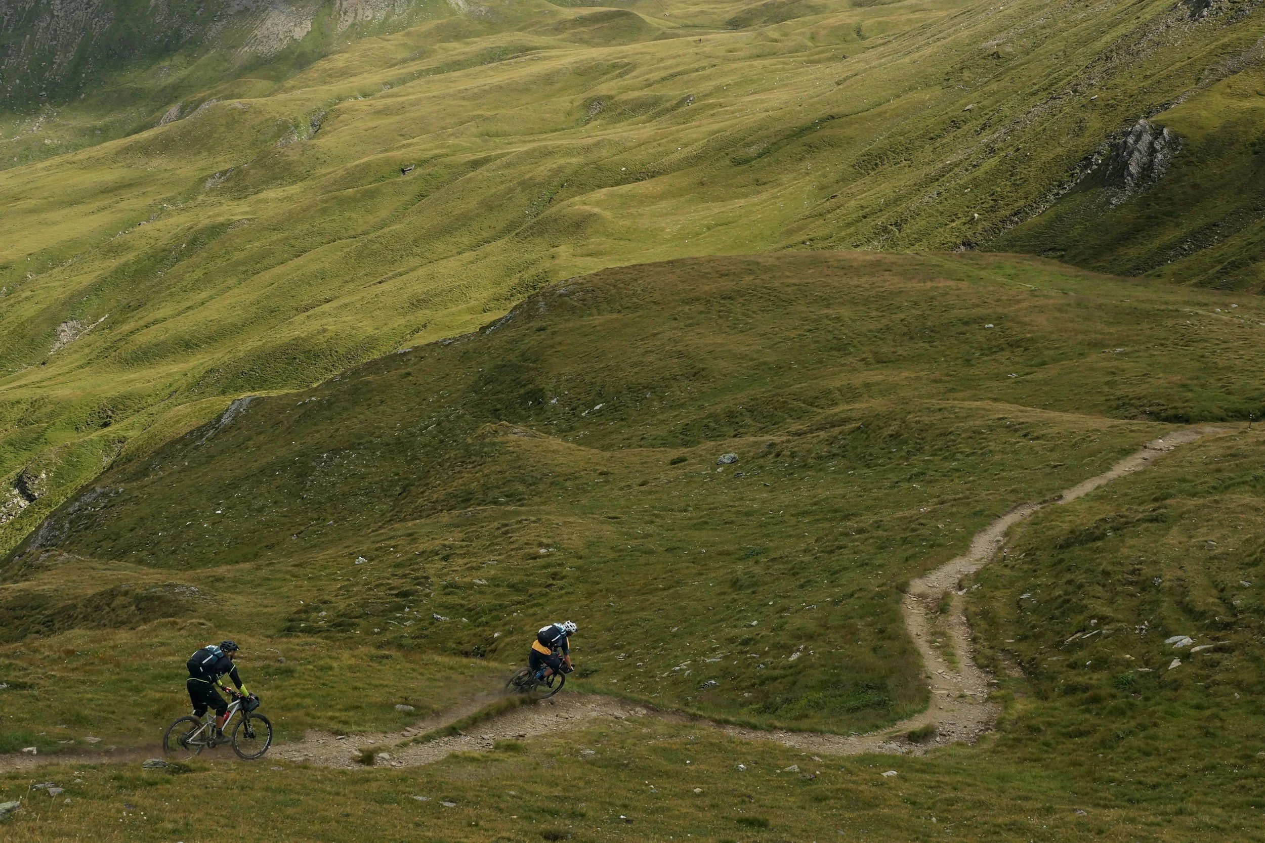 Two cyclists riding mountain bikes on a dirt trail through green hills under cloudy sky.
