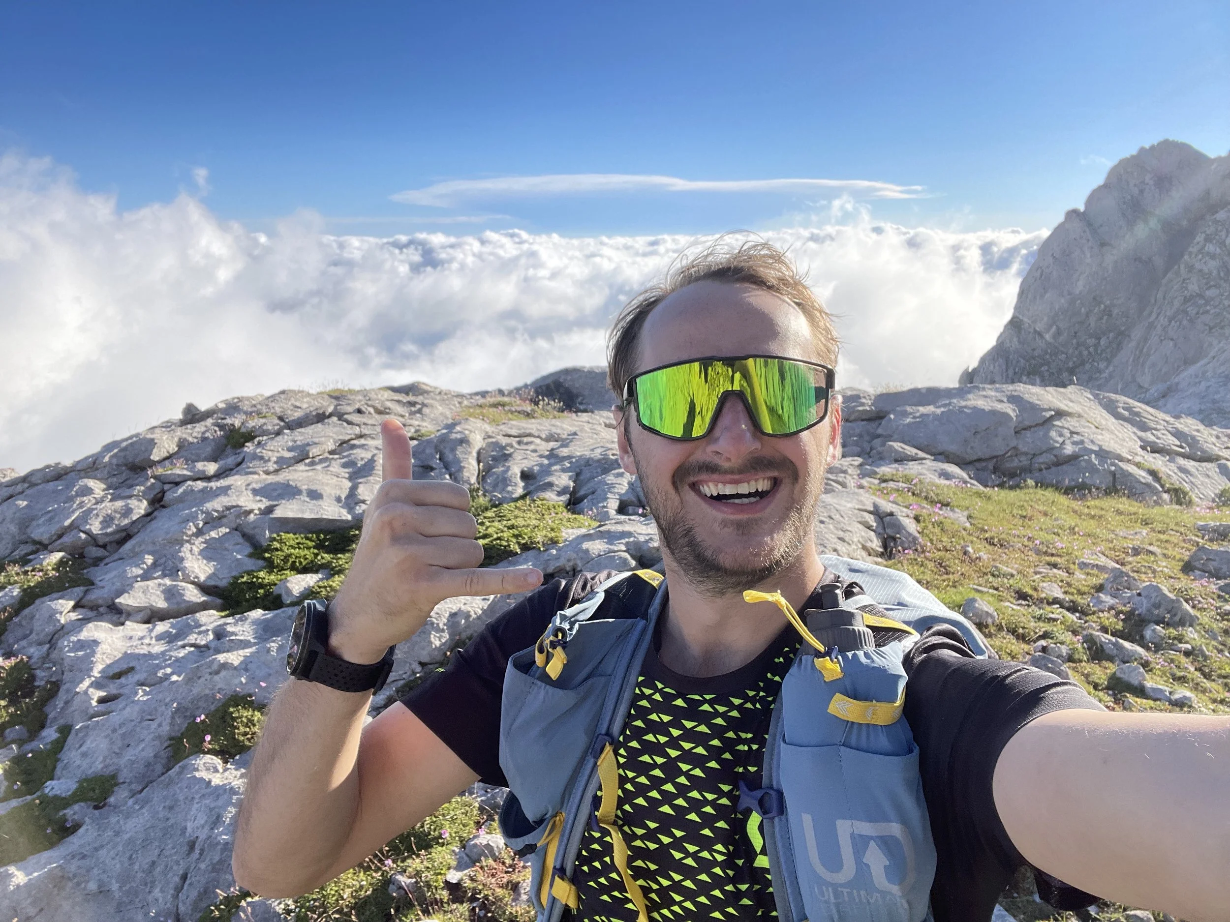 A man taking a selfie on a mountain trail with rocks and green grass, smiling, wearing sunglasses, a black shirt with yellow patterns, and a backpack, with a scenic cloudy sky background.