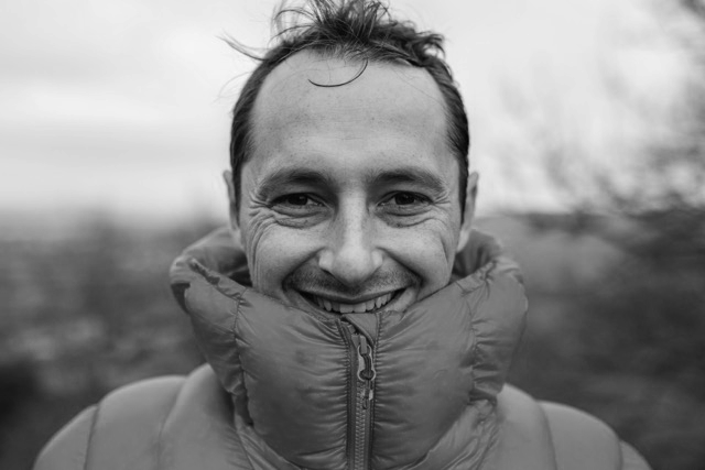 Black and white photo of a smiling man outdoors, wearing a puffy jacket zipped up to his chin.