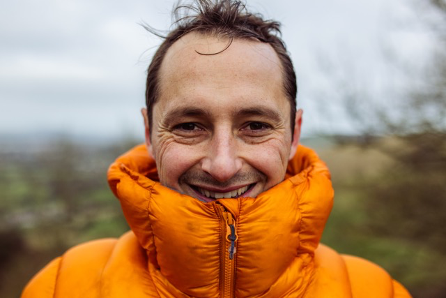 Man smiling outdoors wearing an orange jacket with the collar zipped up.