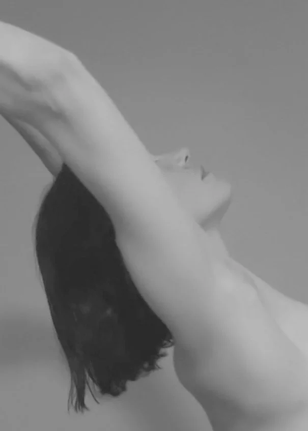 Black and white photo of Kat Slade with dark hair, eyes closed, arms raised above her head, in a relaxed pose.