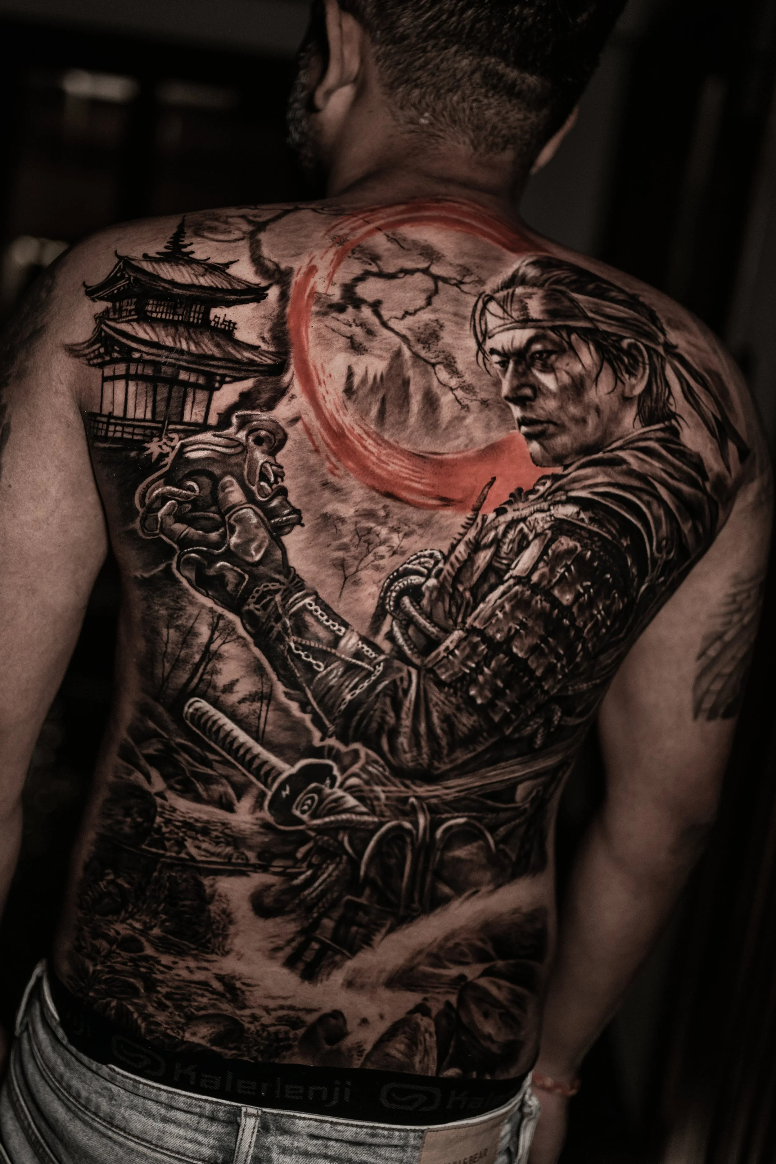 A detailed black and gray tattoo on a man's back depicting a samurai warrior, a Japanese pagoda, mountains, and a red crescent moon.