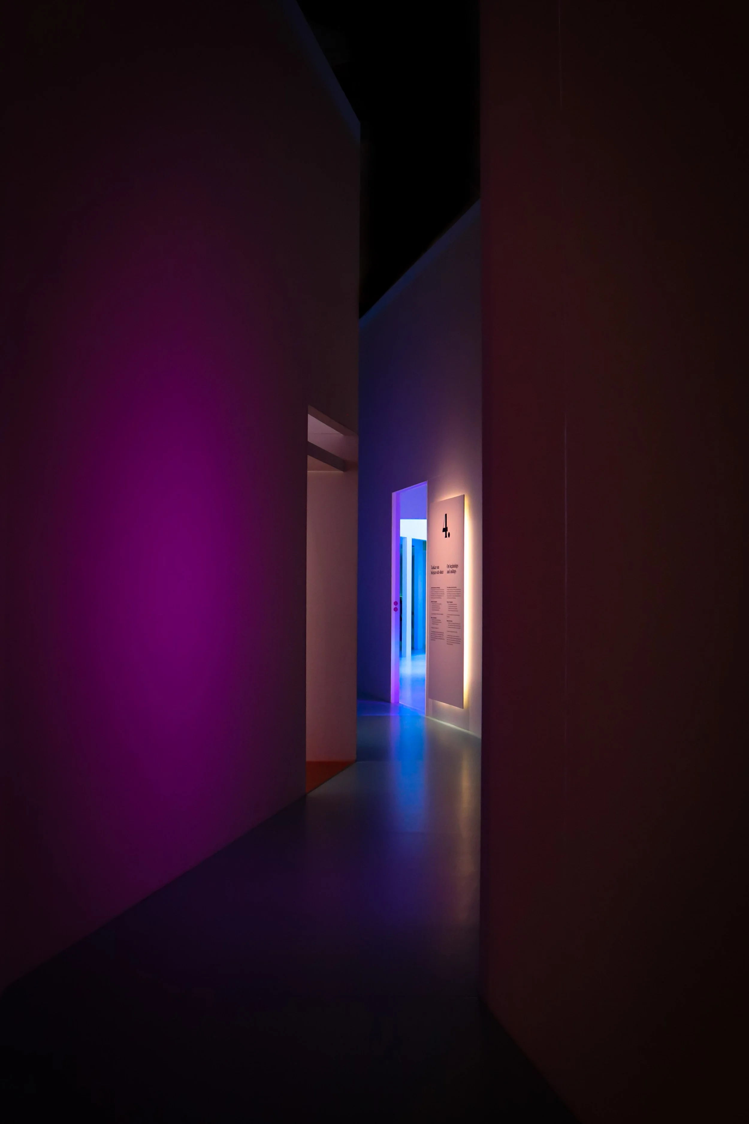 Couloir sombre éclairé par des lumières colorées, avec des murs violets à gauche et bleus à l'arrière-plan. Un panneau blanc est visible sur le mur à droite.