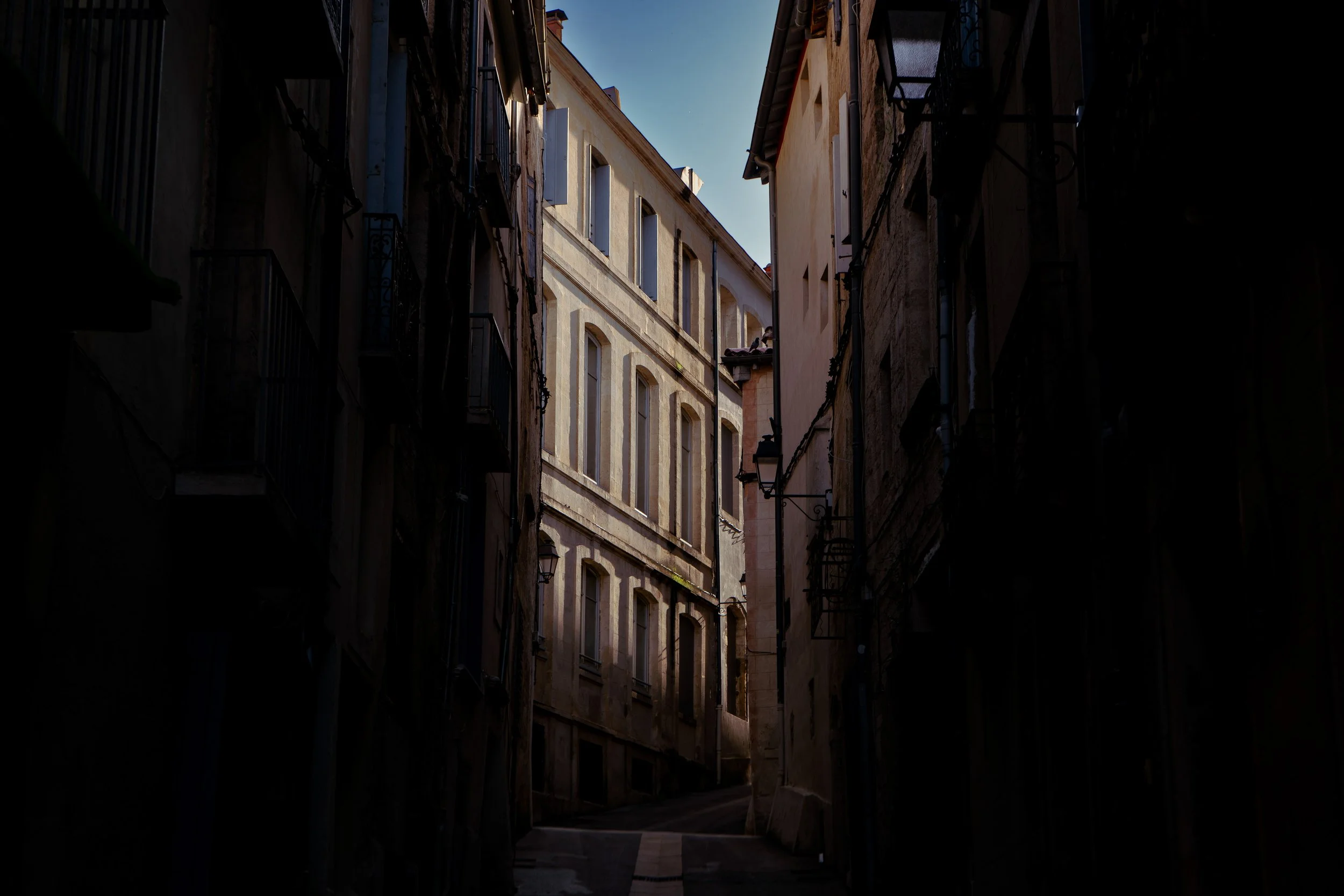 Rue étroite avec bâtiments anciens éclairés par le soleil, façades en pierre.