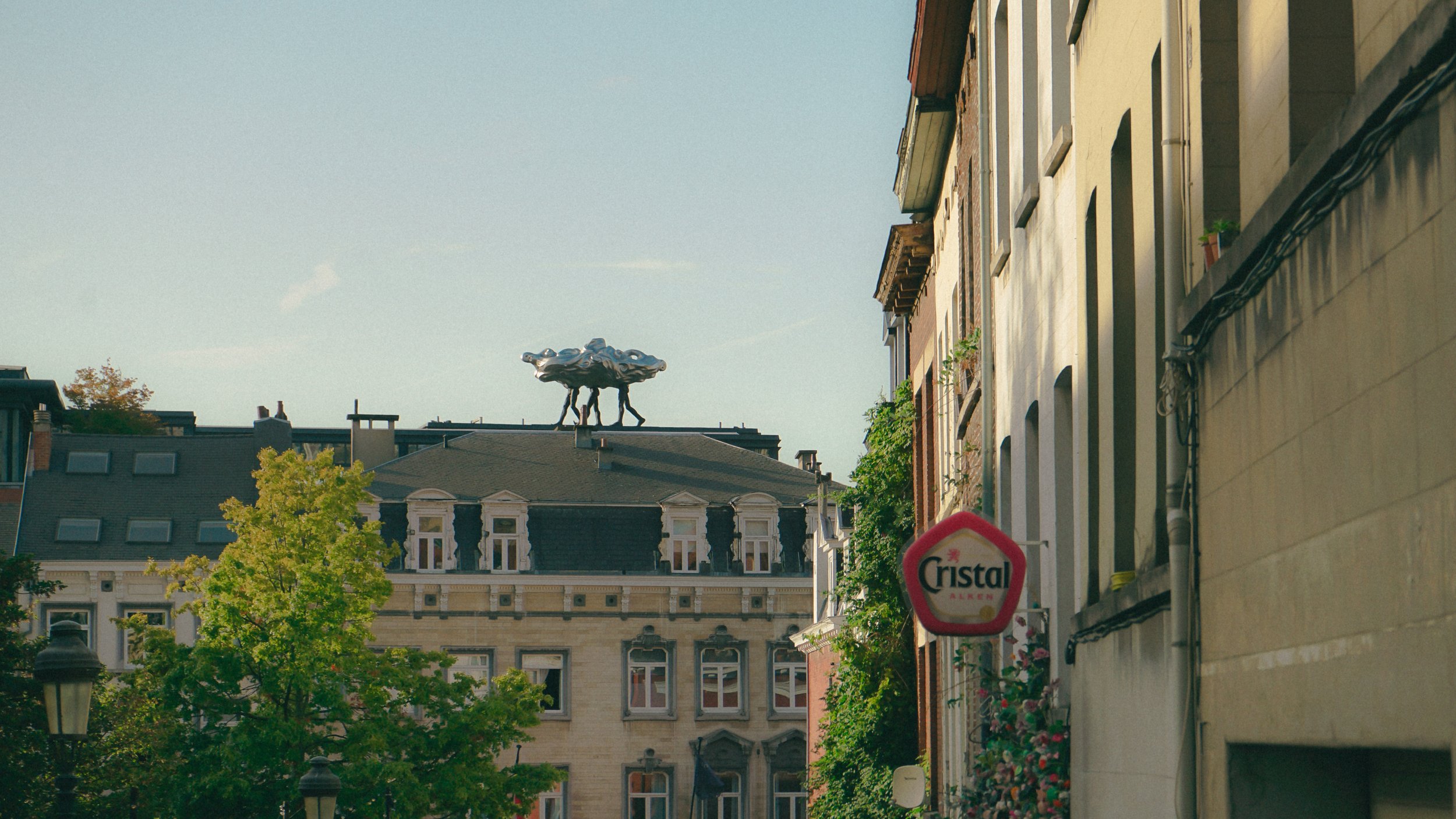 Rue urbaine avec des bâtiments anciens, un arbre, un panneau 'Cristal Alken', et une sculpture sur un toit.
