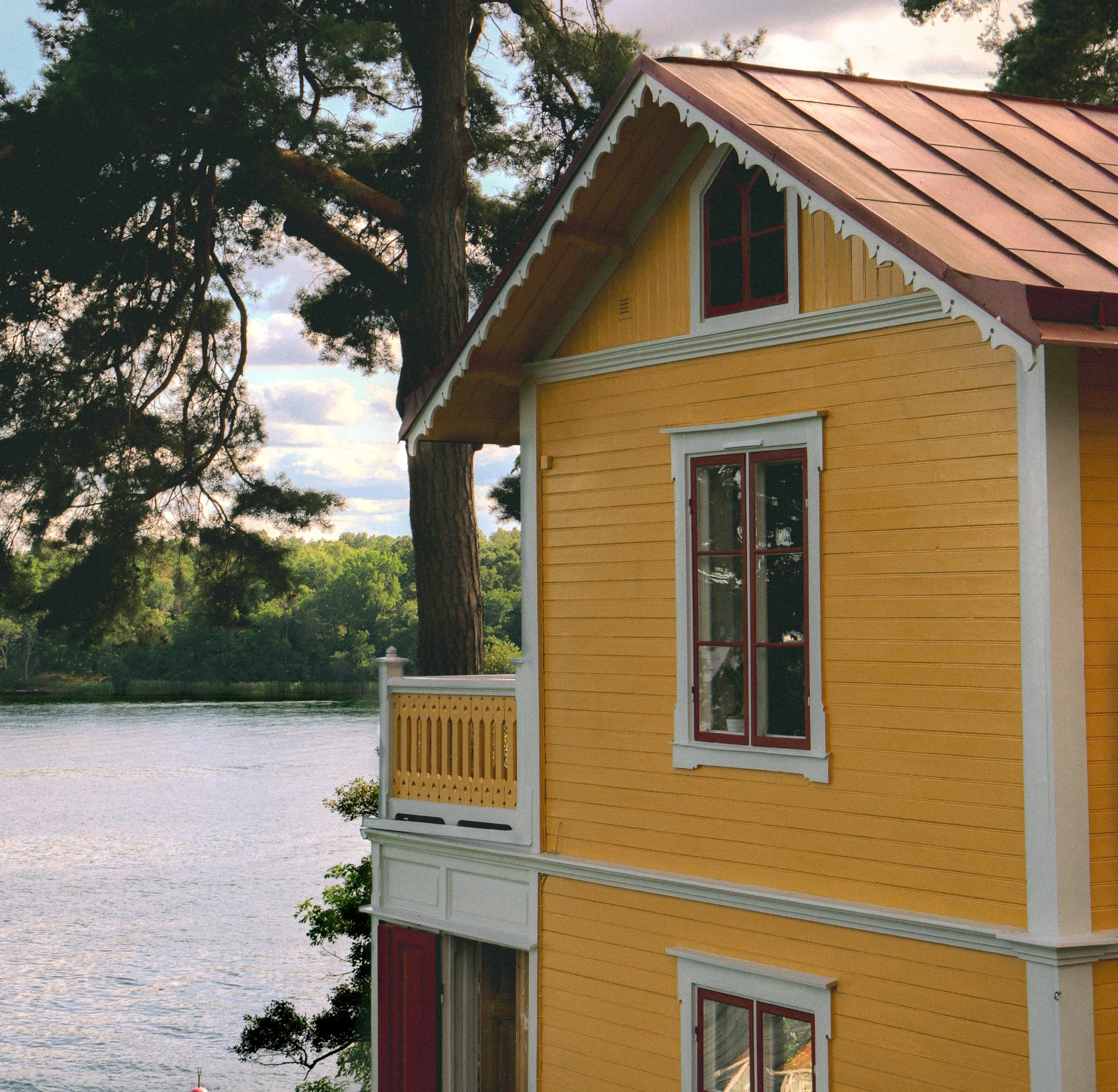 Façade d'une maison jaune avec un toit en métal rouge, située près d'un lac entouré d'arbres.