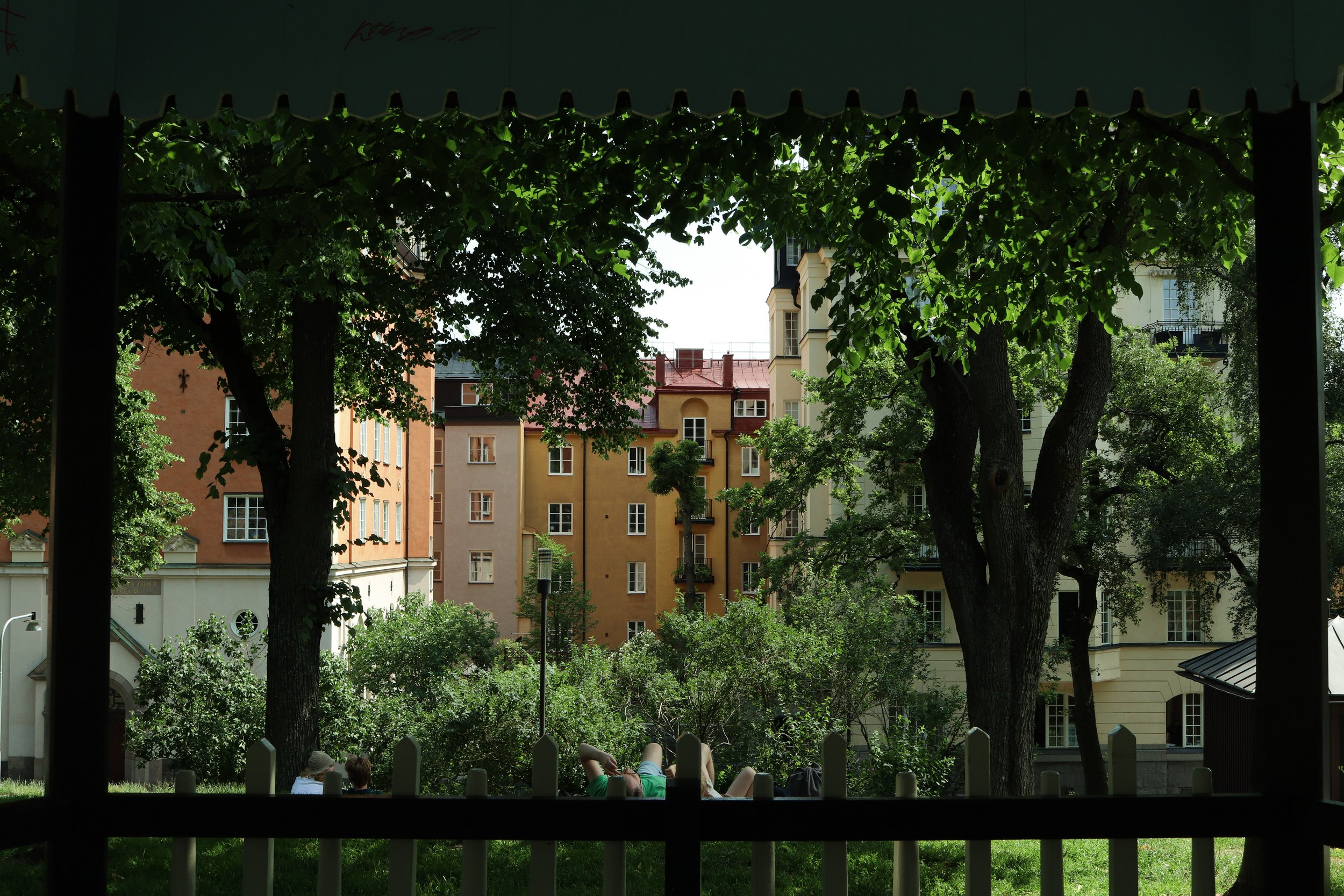 Vue de bâtiments colorés à travers des arbres depuis un espace ombragé avec une clôture blanche au premier plan. Des personnes se reposent dans l'herbe.