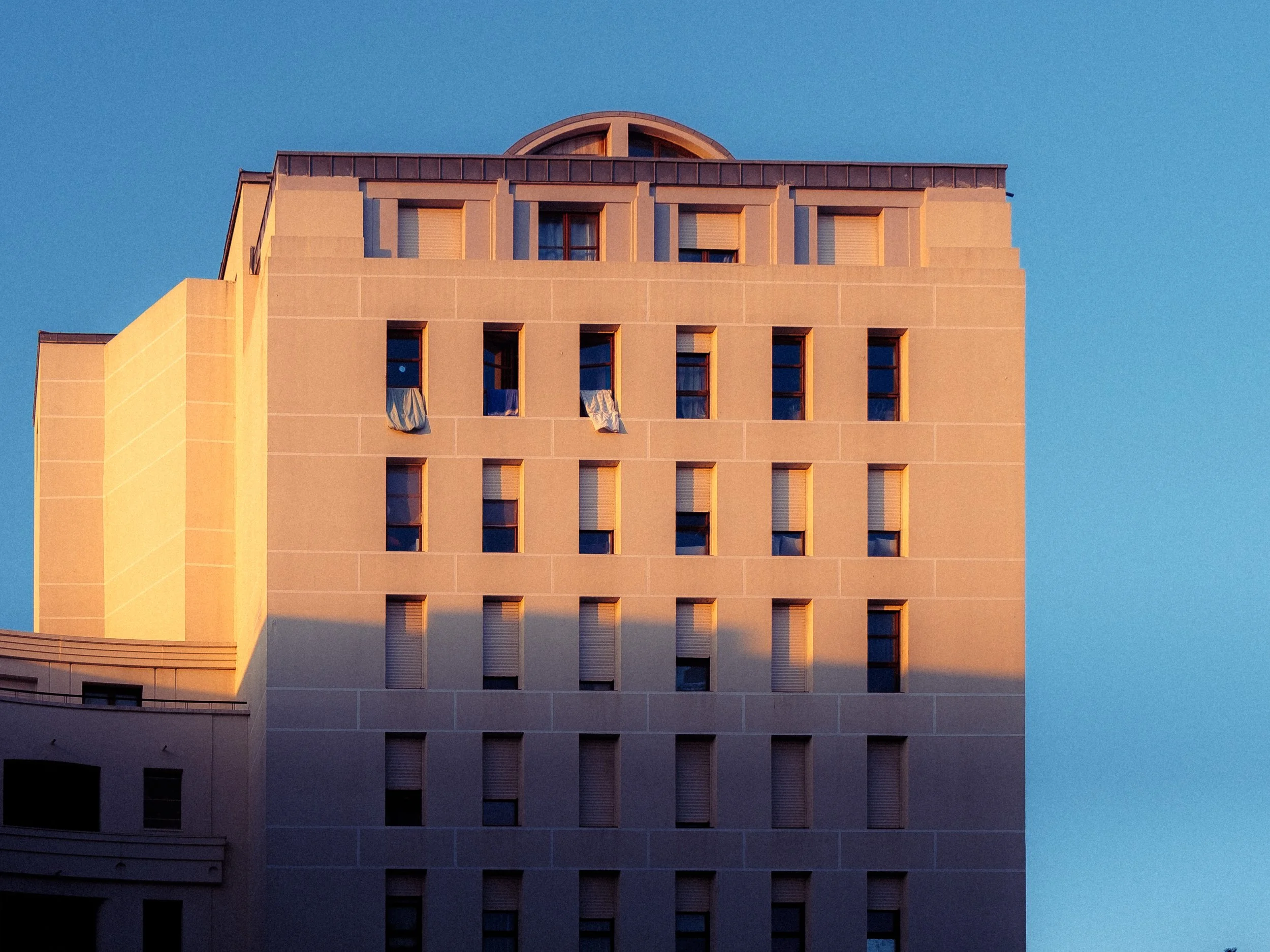 Bâtiment beige moderne éclairé par le soleil couchant, avec des fenêtres carrées et un ciel bleu clair.