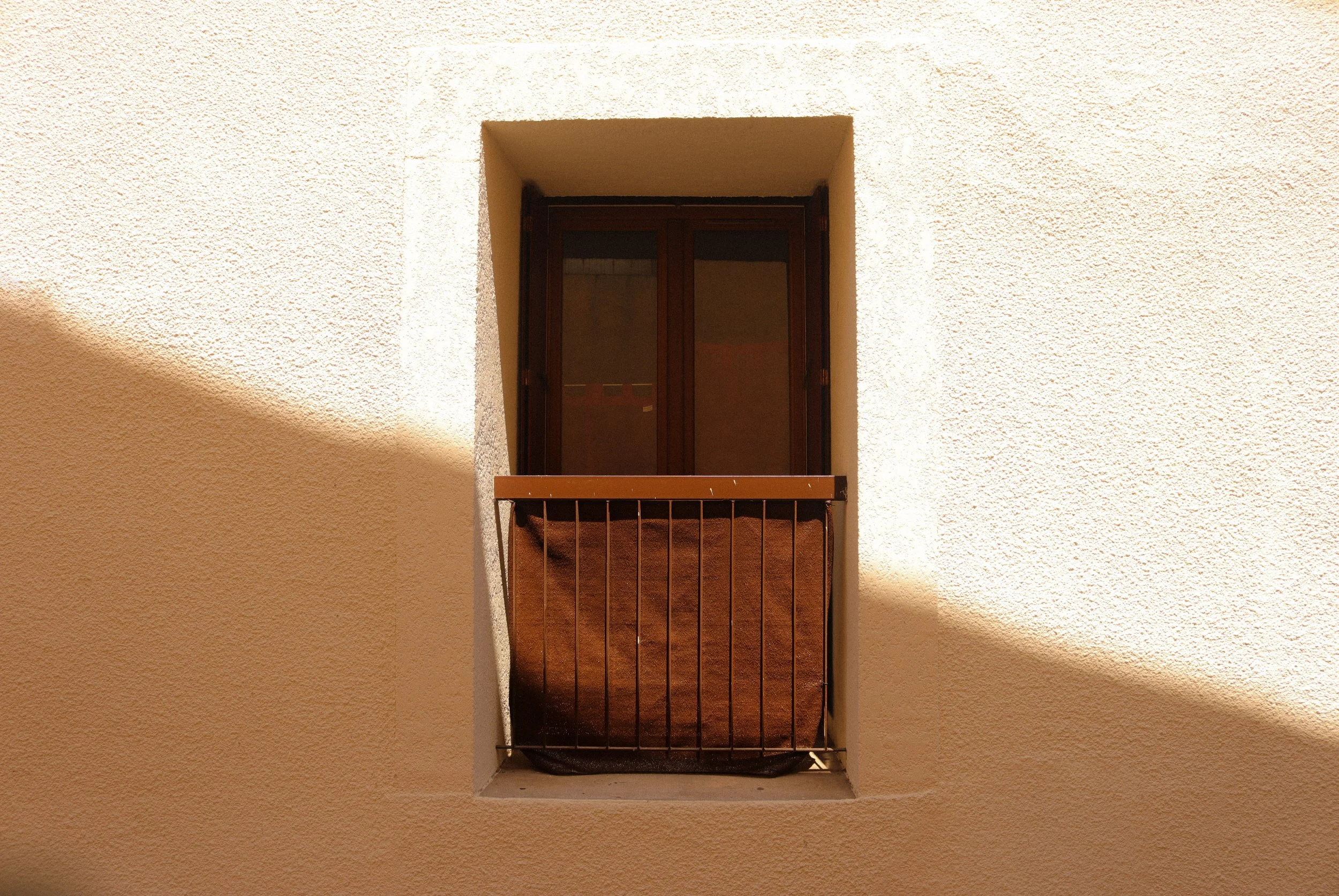 Fenêtre encadrée de plâtre beige avec rambarde en métal et ombre diagonale sur le mur.
