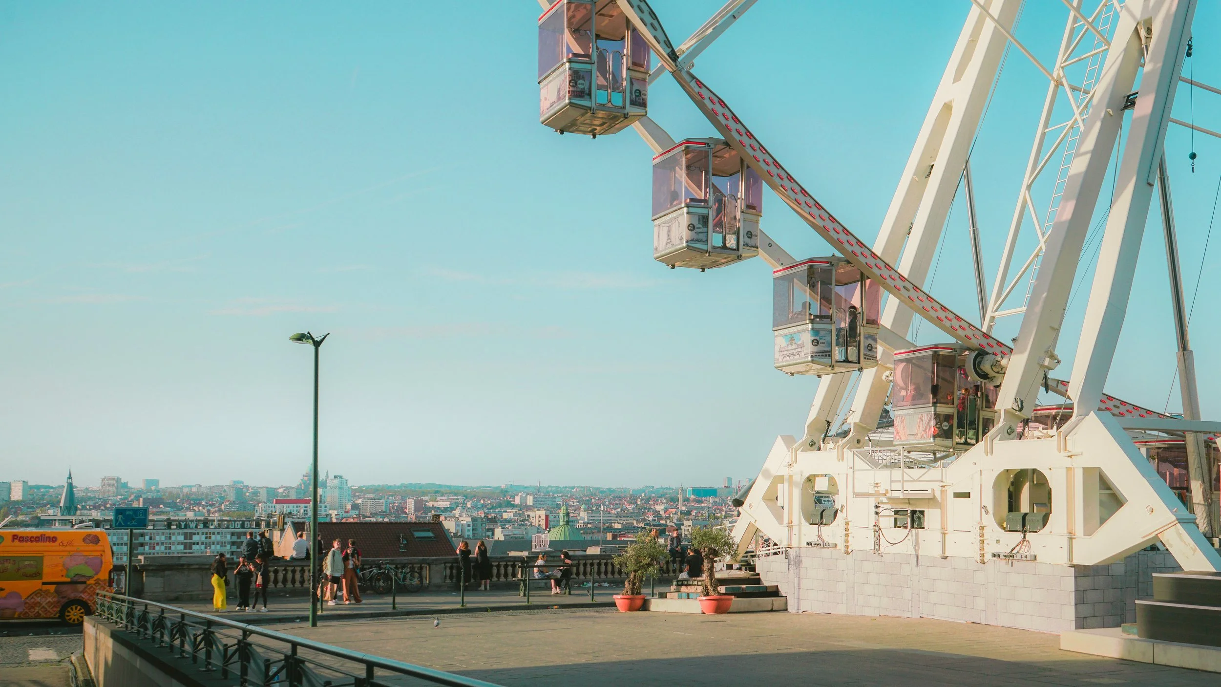 Grande roue avec des cabines surplombant une ville, ciel bleu en arrière-plan, quelques personnes debout sur une terrasse.