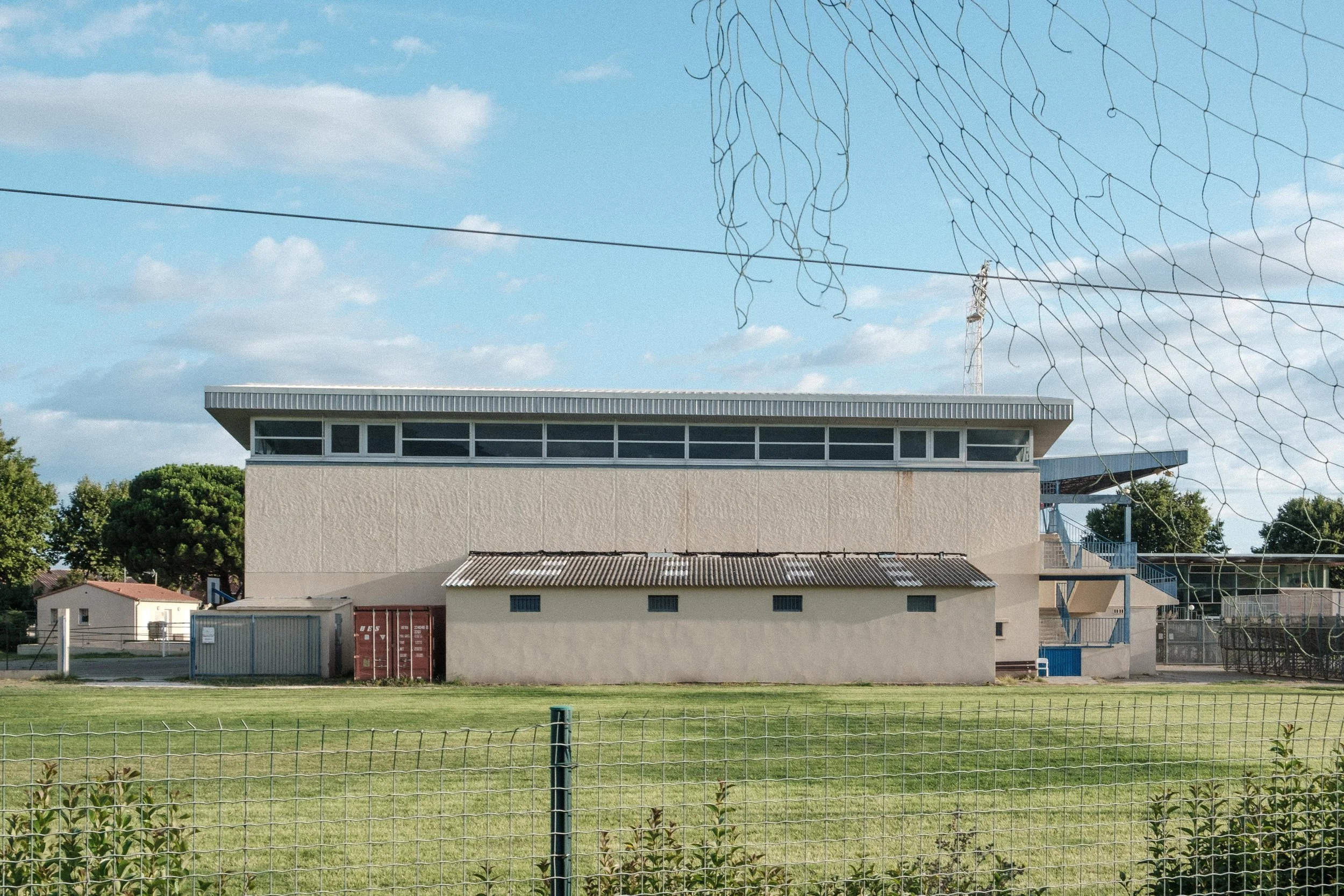 Bâtiment sportif avec fenêtre en haut et terrain en avant, entouré d'une clôture et de végétation, ciel partiellement nuageux