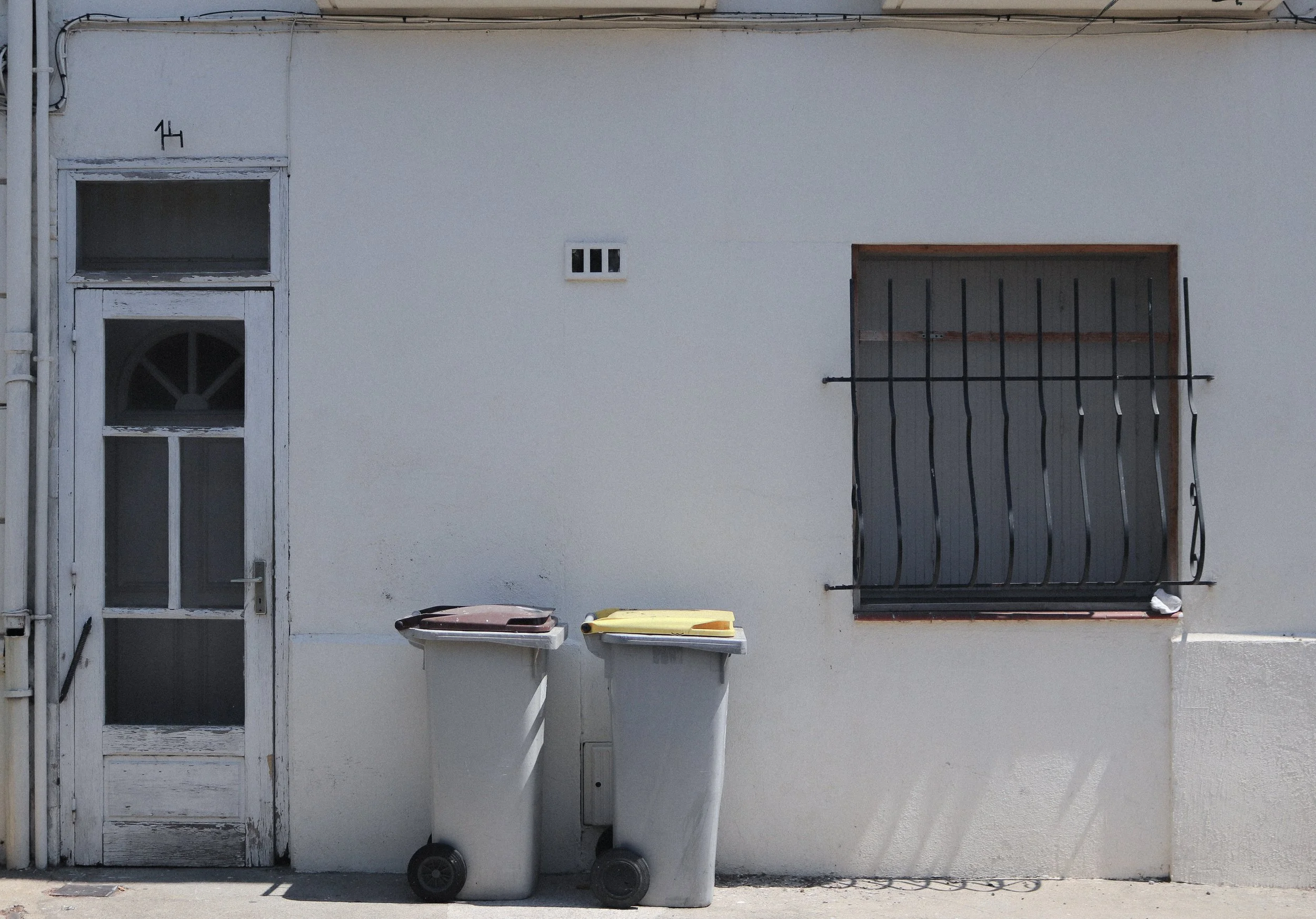 Façade d'une maison avec porte, fenêtre grillagée et deux bacs à ordures.