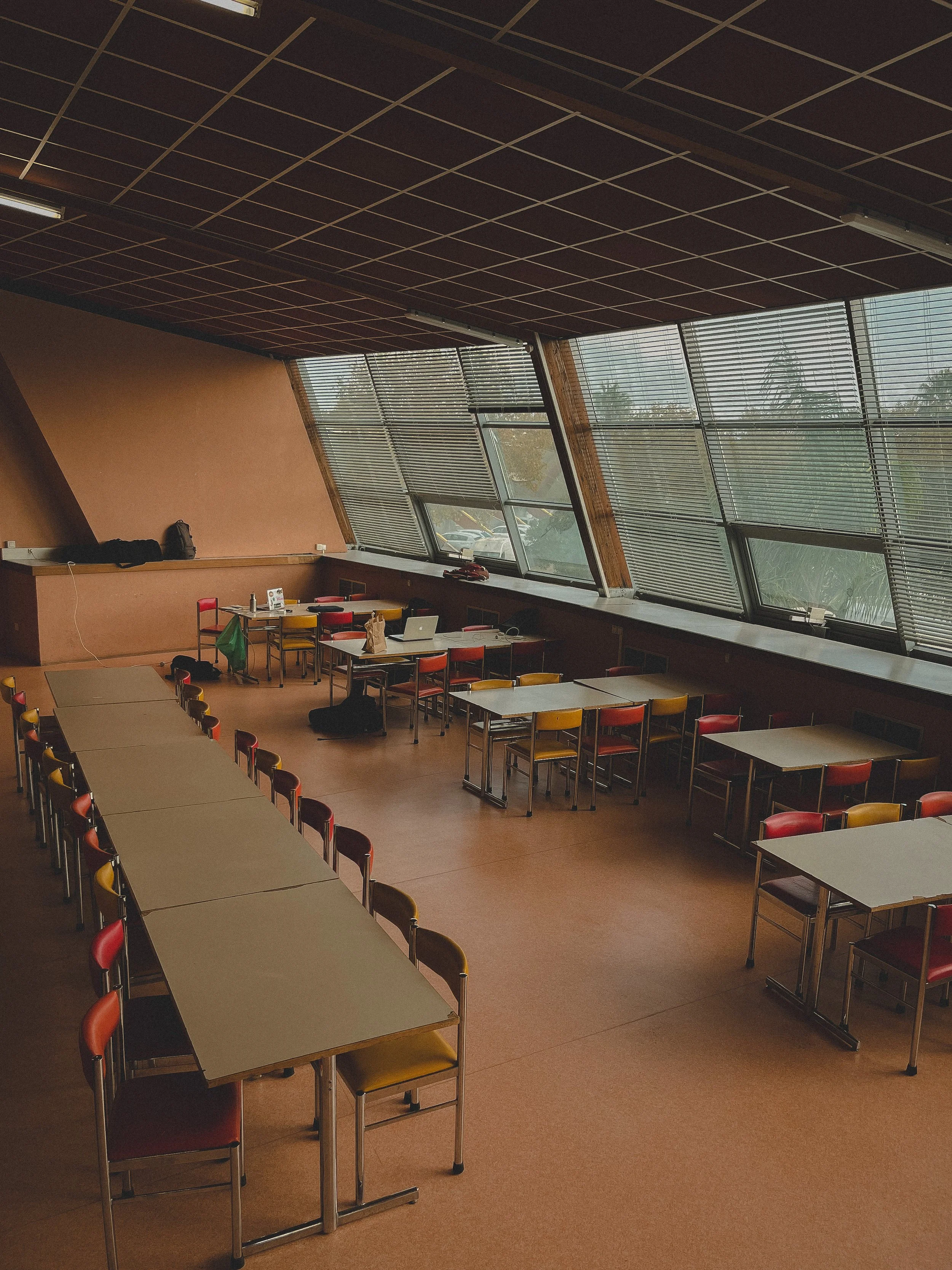 Une salle de classe vide avec des tables et des chaises colorées près de fenêtres inclinées.