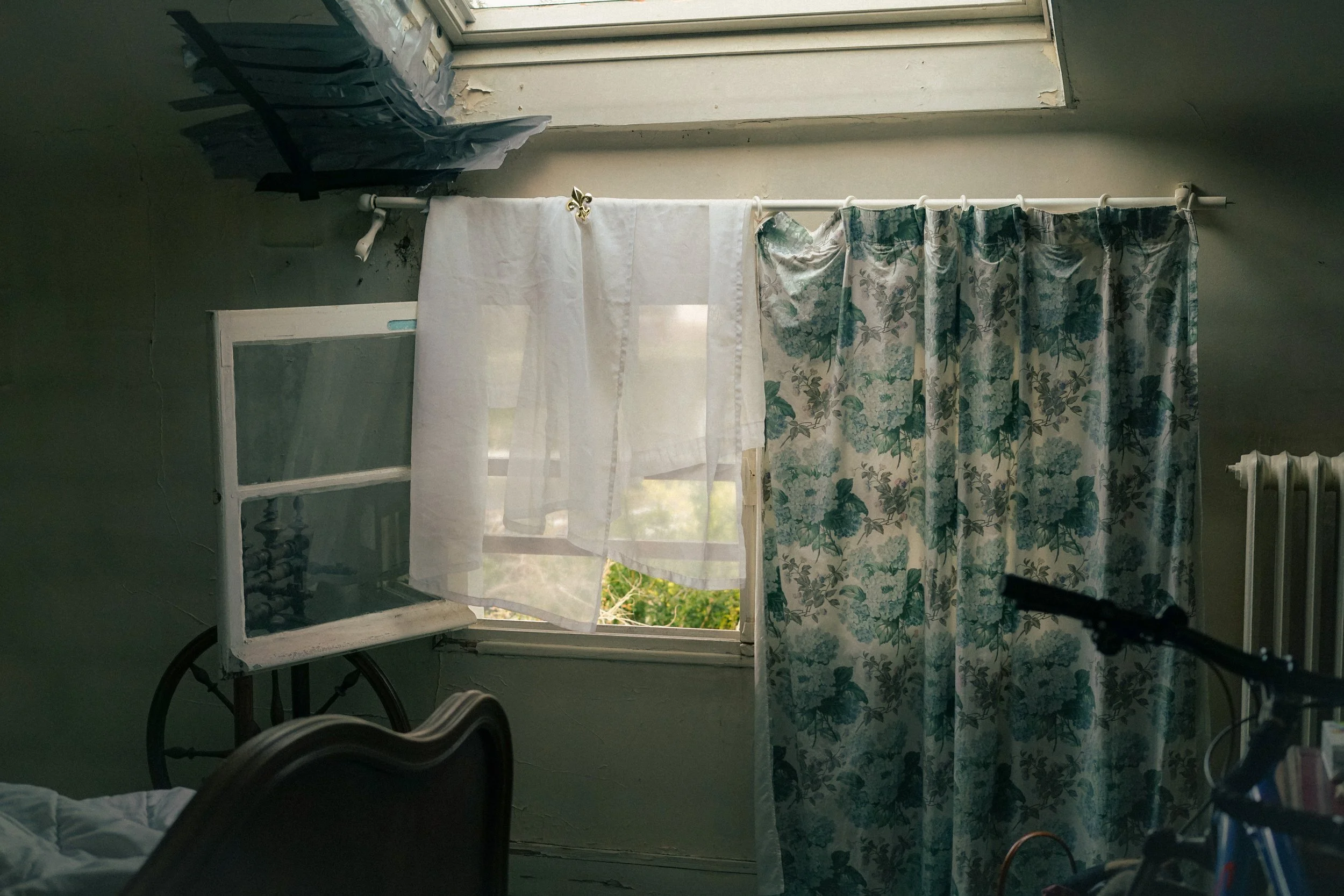 Intérieur d'une chambre avec une fenêtre ouverte, des rideaux floraux, un lit partiellement visible et un radiateur.