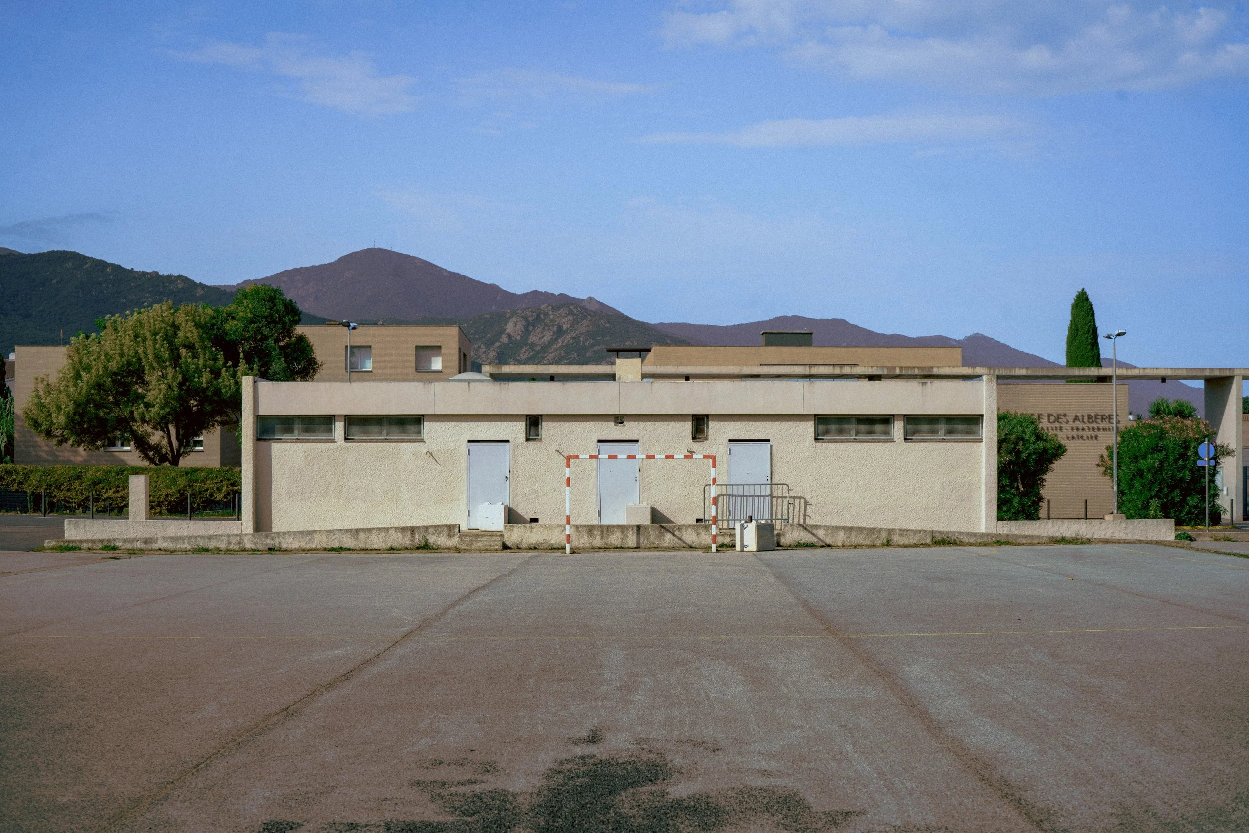 Bâtiment moderne beige avec des portes et fenêtres, terrain devant, montagnes et ciel bleu en arrière-plan.
