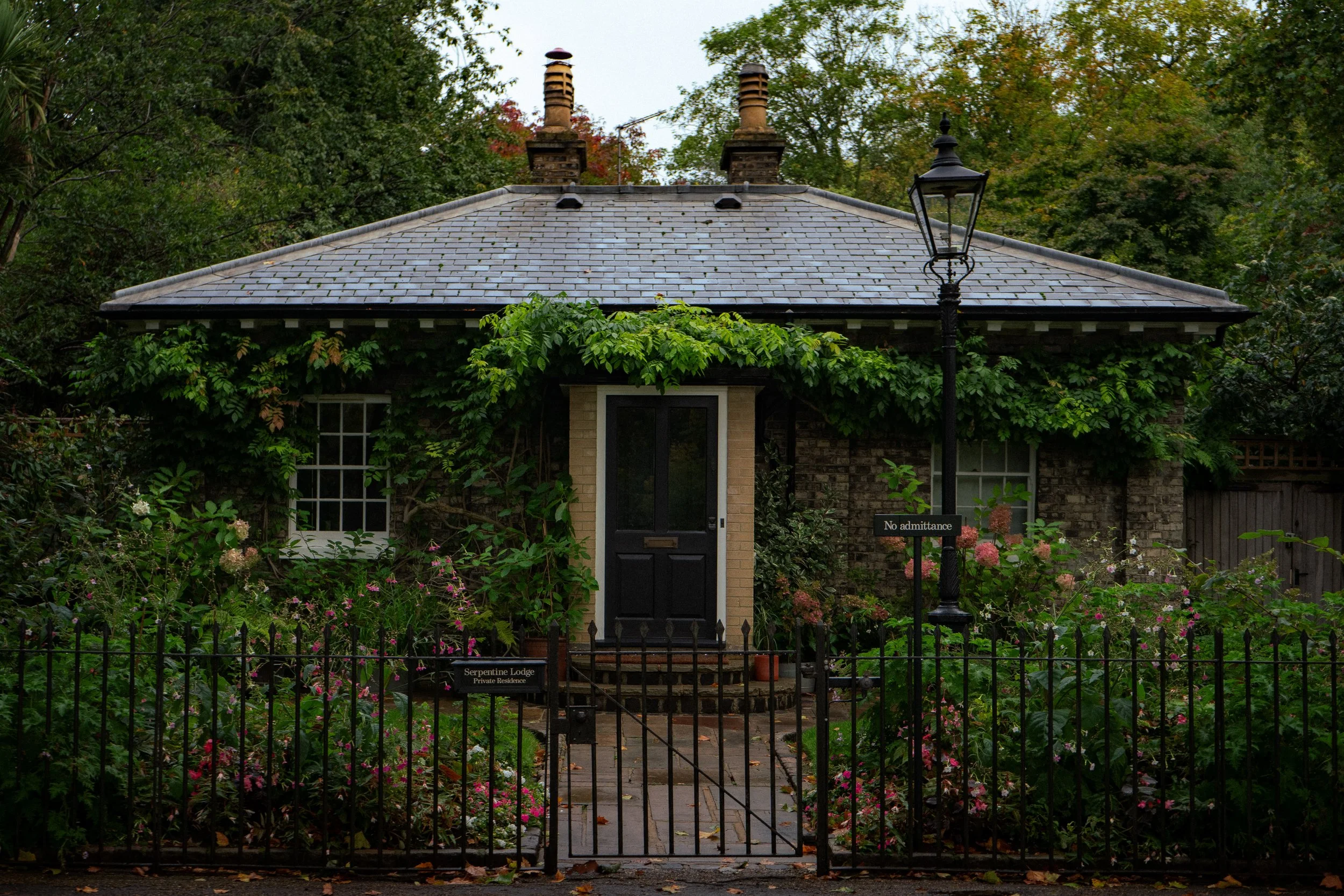Petite maison couverte de lierre avec toit en ardoise, entourée de jardins fleuris et clôturée par une grille en fer, avec un lampadaire à l'extérieur.