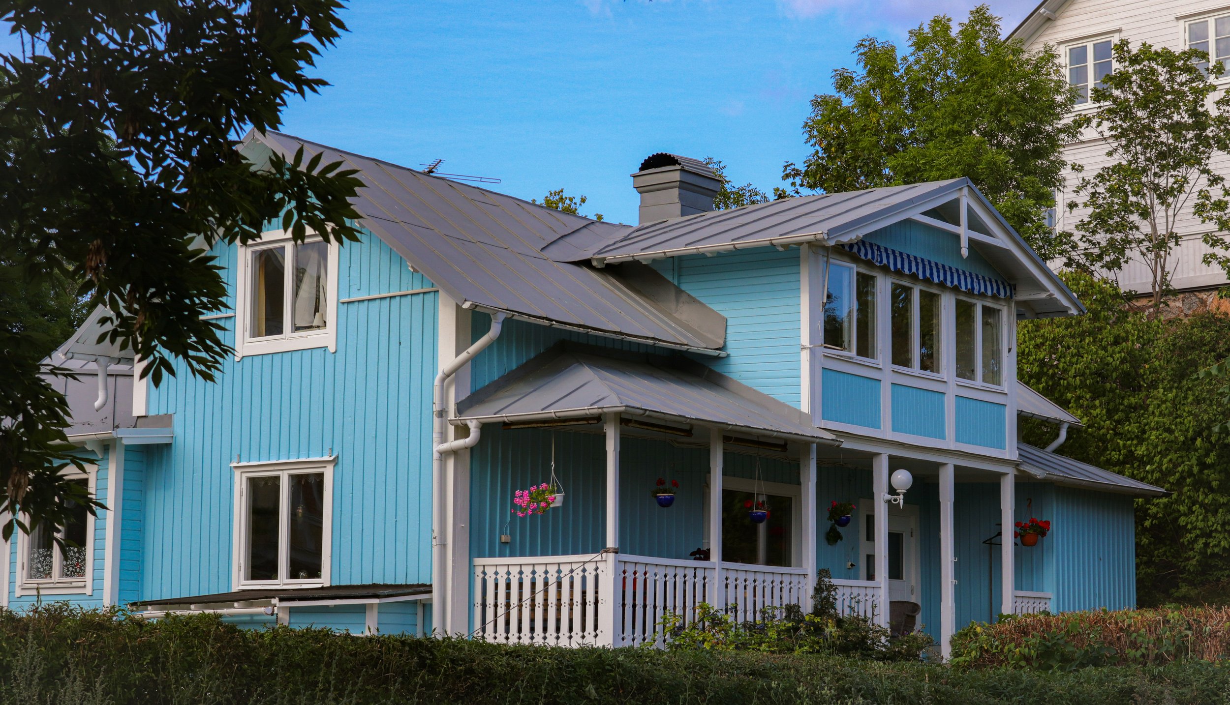 Maison en bois bleue avec jardin et fleurs en pot suspendues, entourée de verdure.