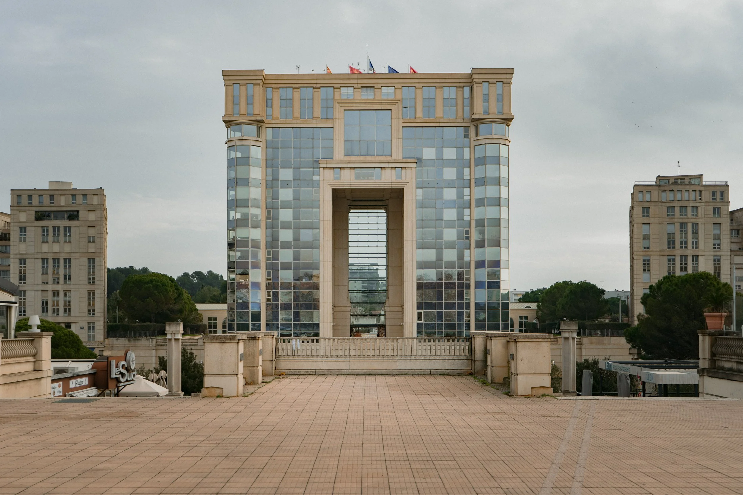 Bâtiment moderne en verre avec trois niveaux principaux. Terrasse pavée au premier plan.
