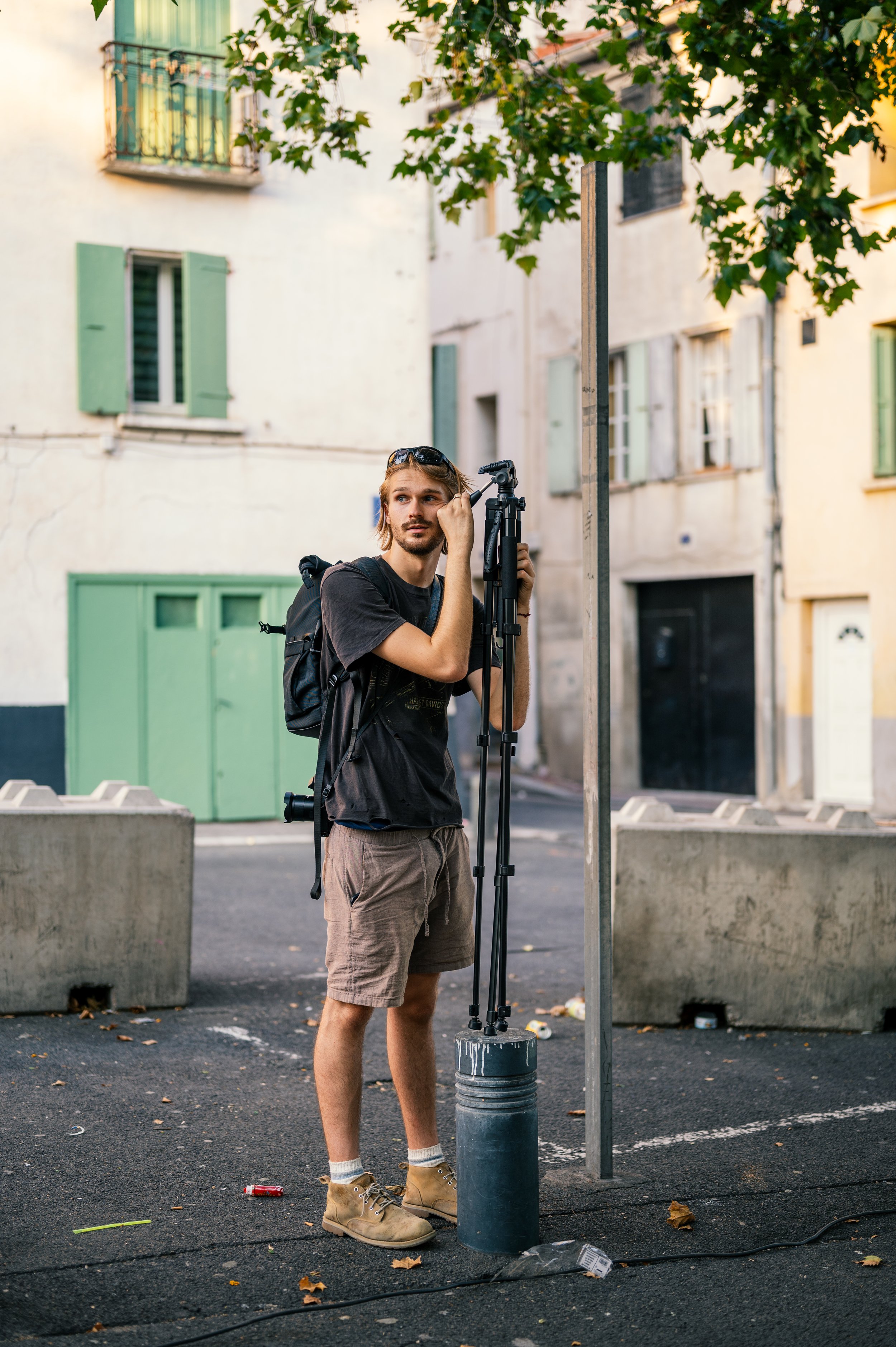 Un jeune homme avec un sac à dos se tient à côté d'un poteau, avec un appareil photo sur un trépied, dans une rue résidentielle.