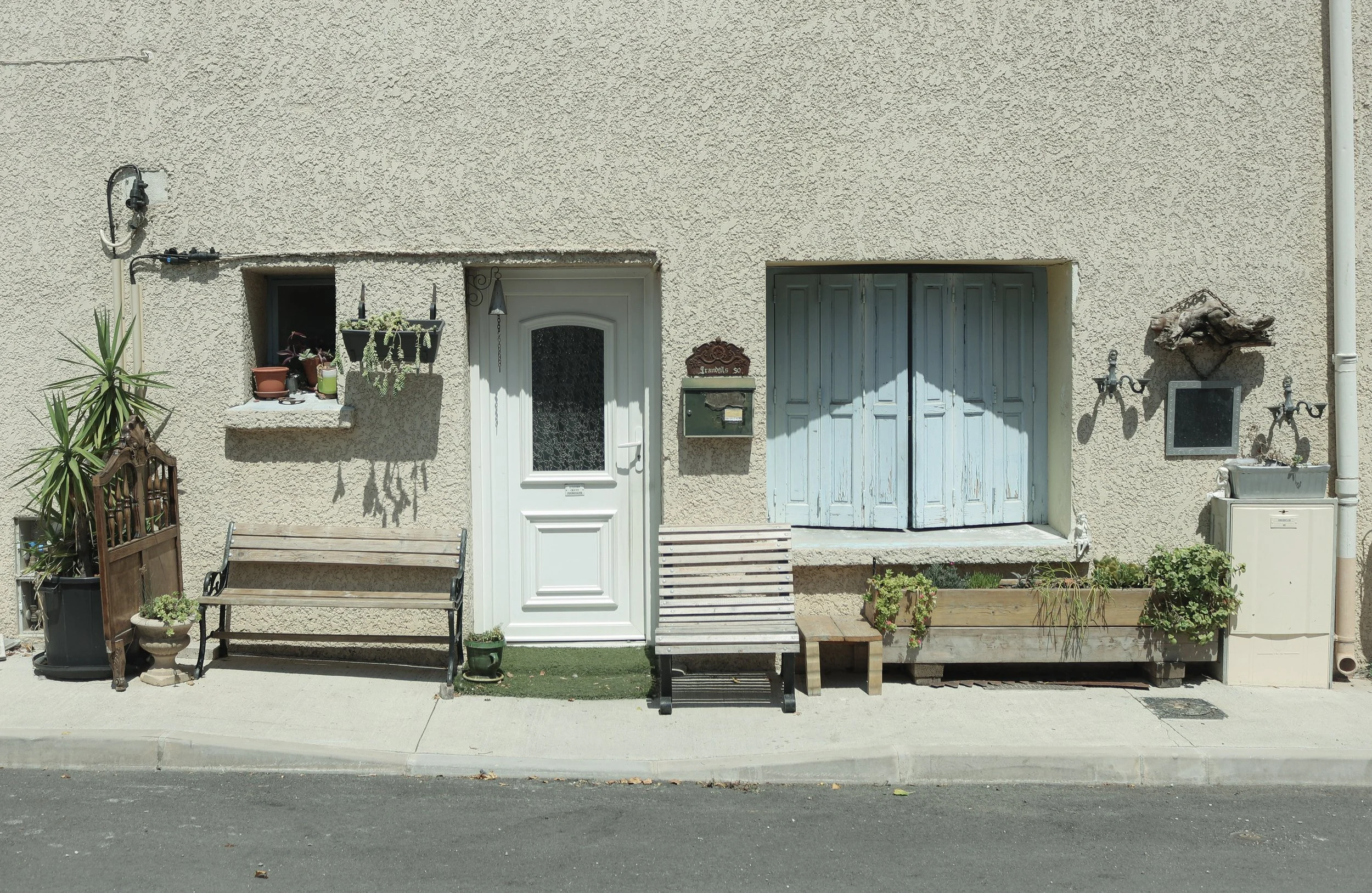 Façade d'une maison avec une porte blanche, des volets bleus fermés, deux bancs en bois, quelques plantes en pot, et des décorations murales.