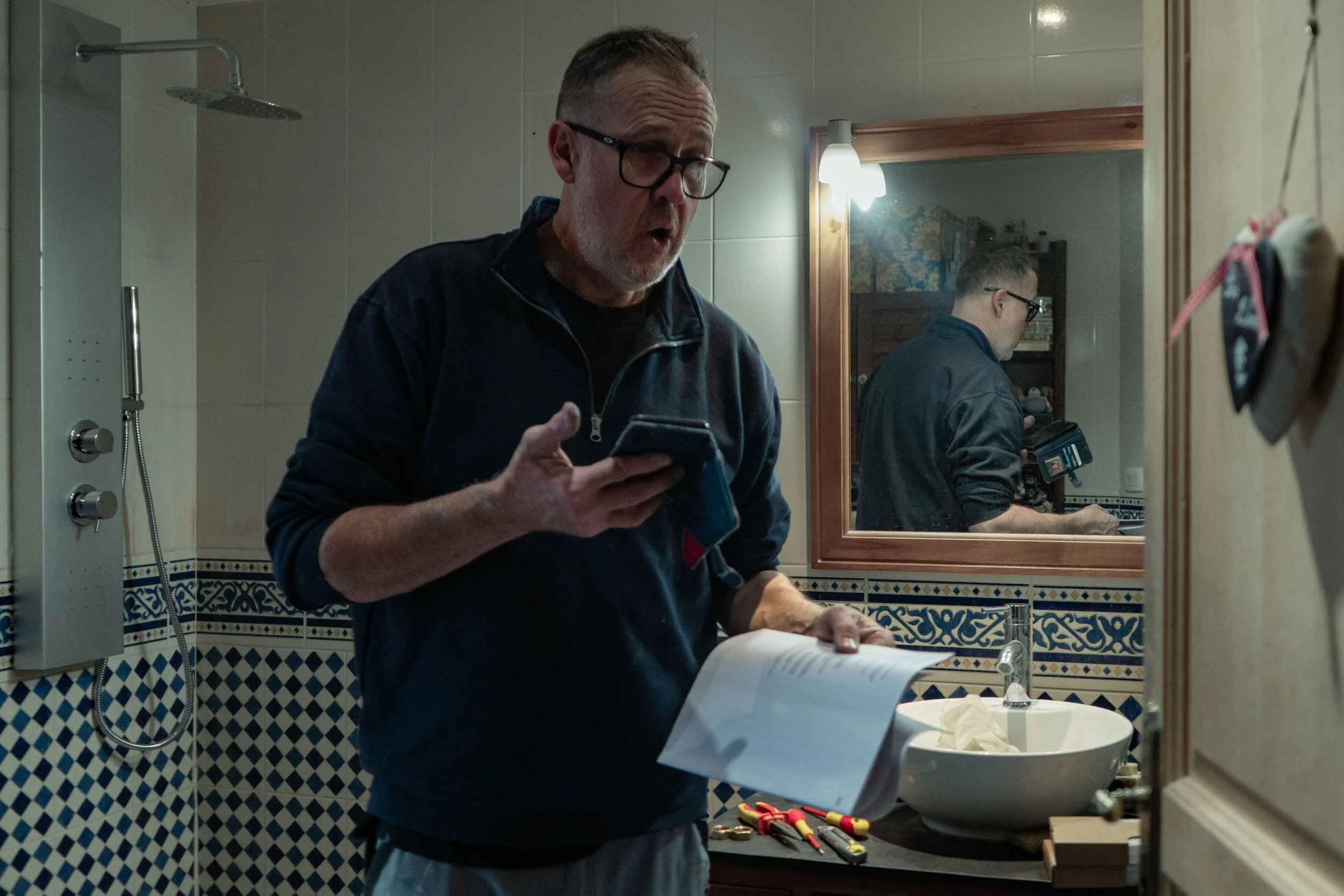 Un homme dans une salle de bain, tenant un smartphone et des documents. Un miroir reflète son image. Un évier et plusieurs outils sont visibles sur le comptoir. Carrelage bleu et blanc en arrière-plan.