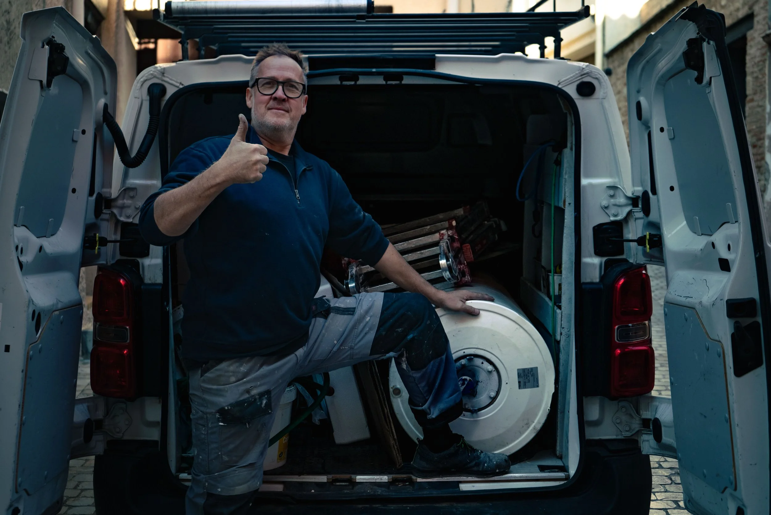 Un homme avec des lunettes fait un geste de pouce levé devant l'arrière ouvert d'une camionnette remplie d'équipement, y compris un gros réservoir métallique.