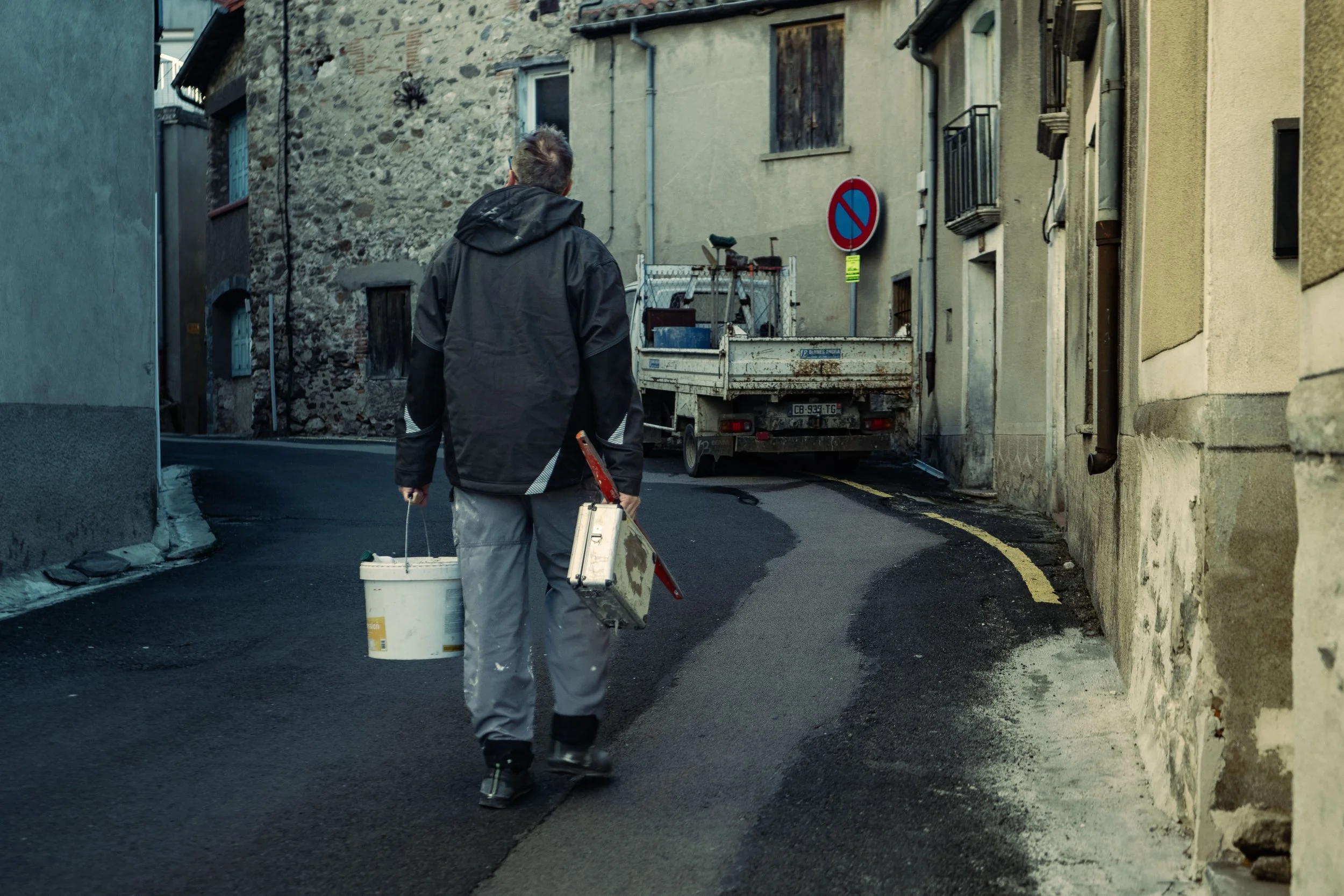 Personne portant un seau et une boîte à outils dans une ruelle étroite, bâtiment en pierre à gauche, camionnette garée plus loin.