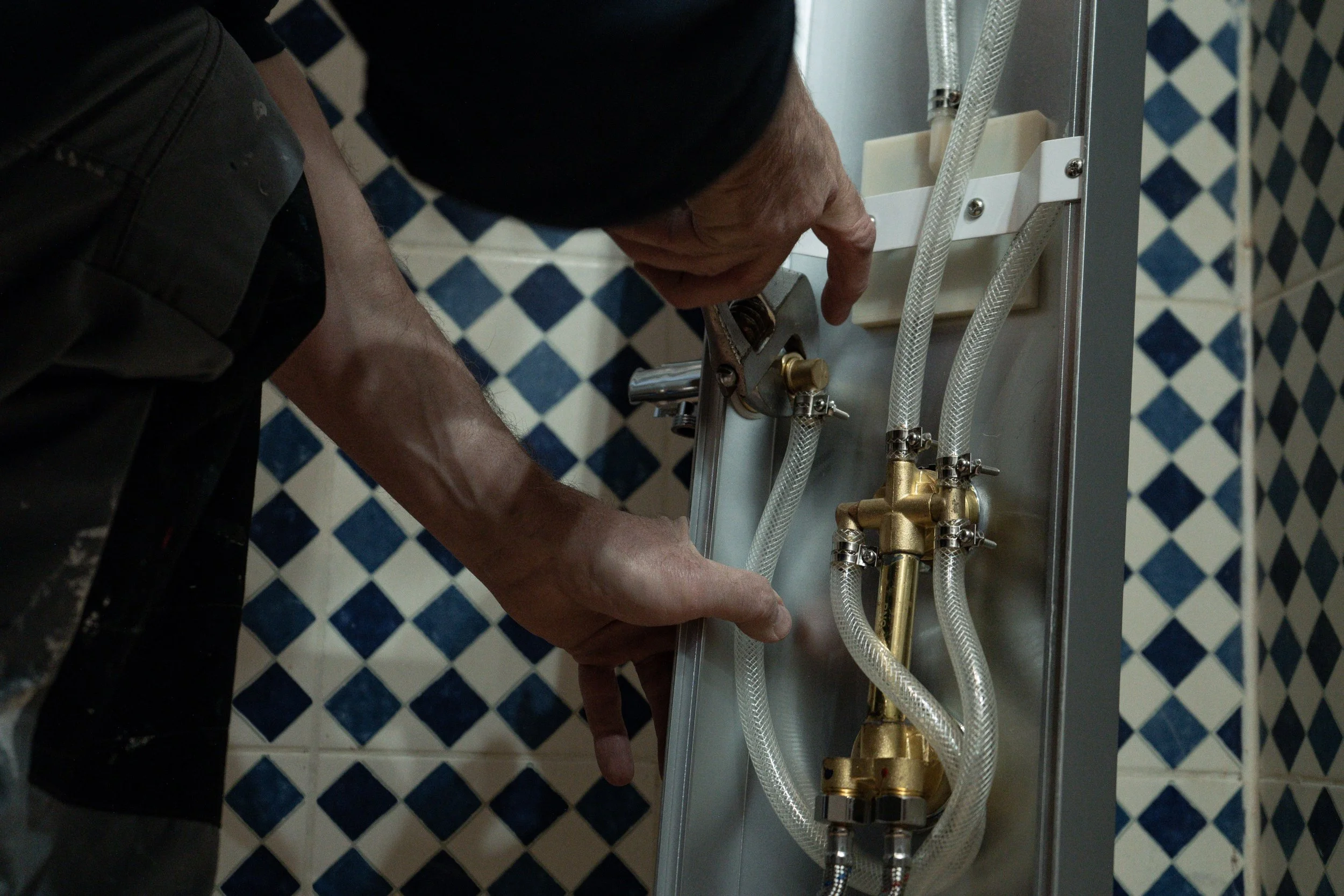 Une personne effectue des réparations de plomberie, ajustant des tuyaux derrière un panneau, avec des carreaux bleus en arrière-plan.