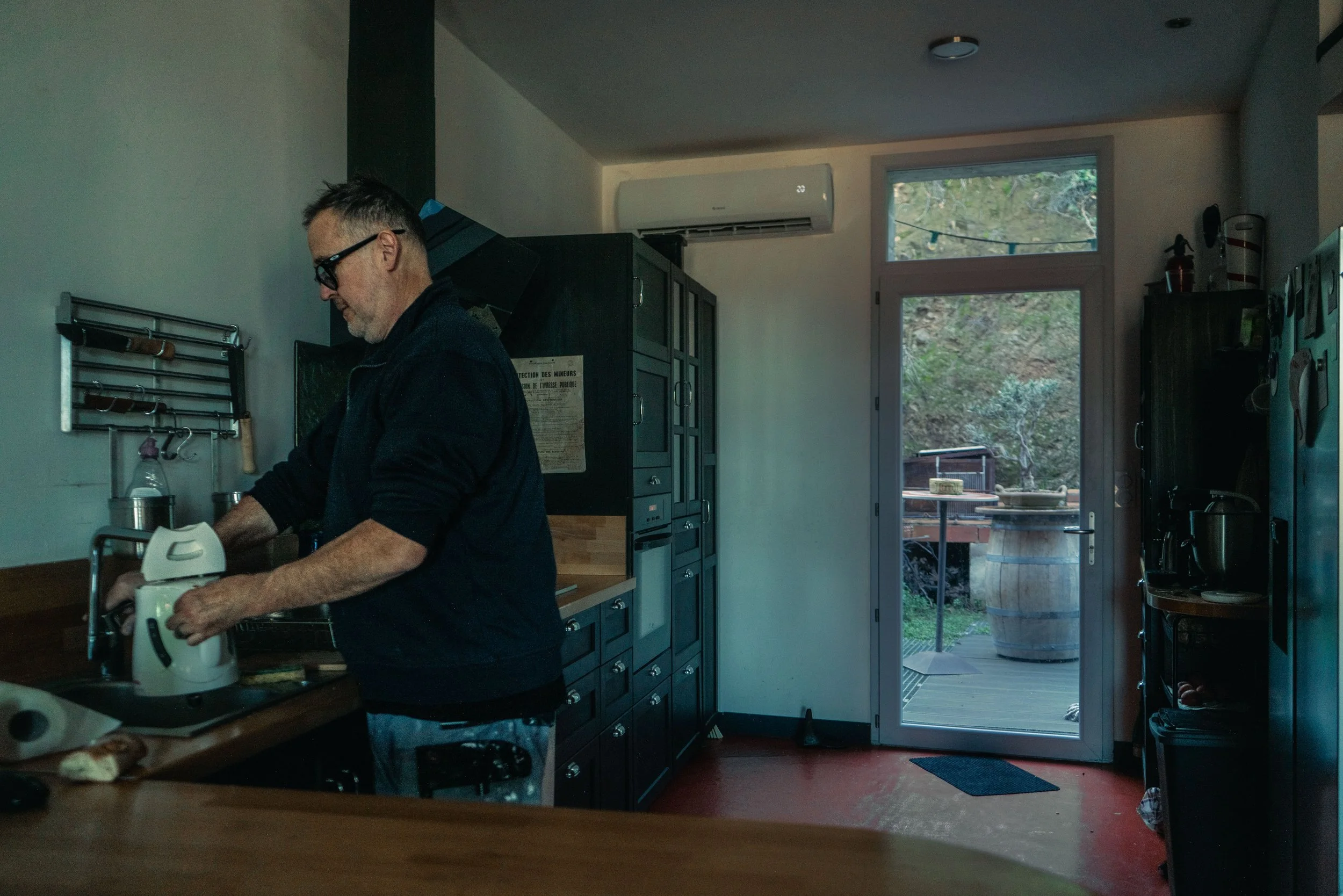 Homme préparant quelque chose dans une cuisine avec des comptoirs en bois, accessoires de cuisine et vue sur un jardin à travers une porte vitrée.